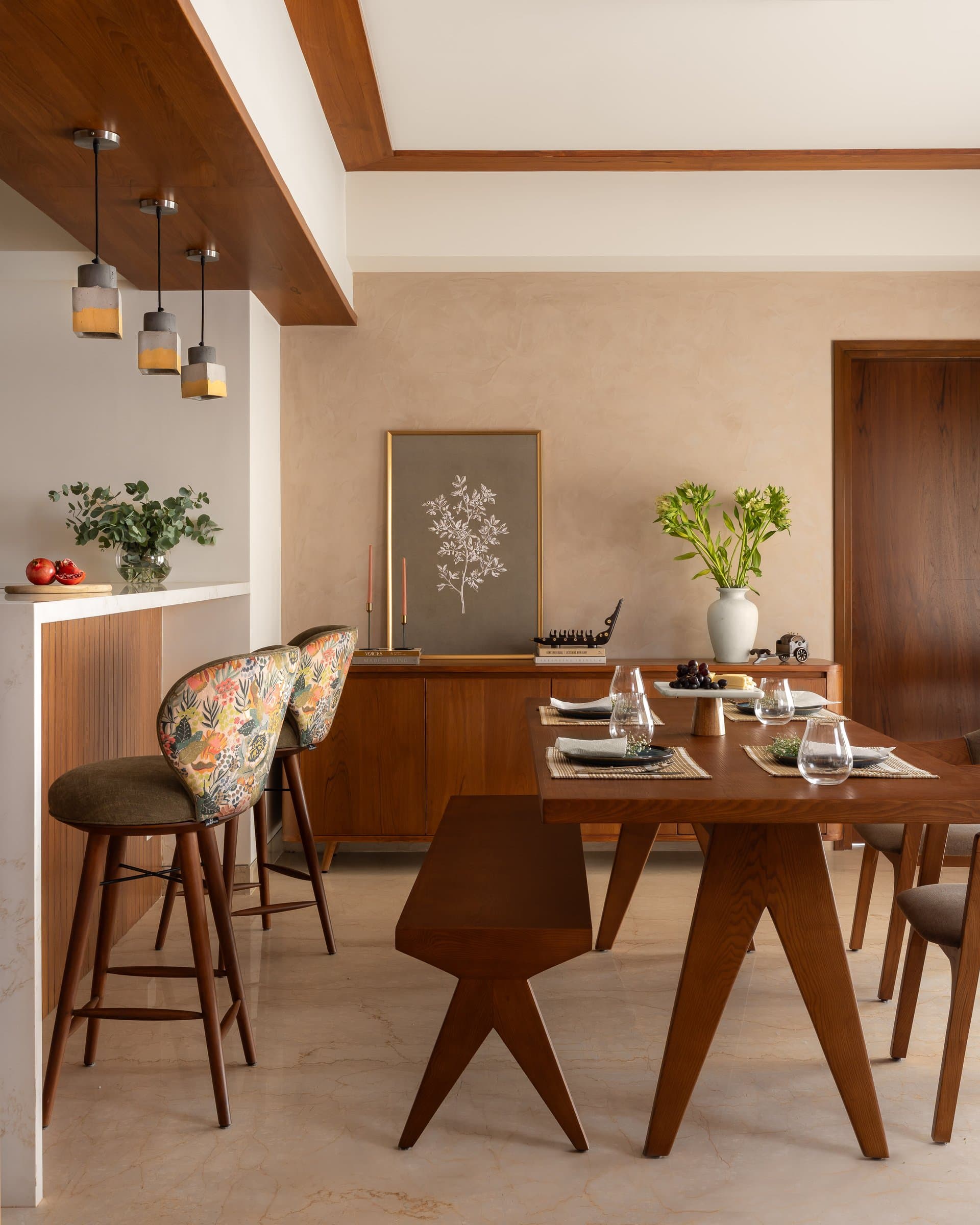 The dining area, with its Chandigarh-referencing timber dining chairs and cluster of concrete pendants anchoring the transition between kitchen counter and table