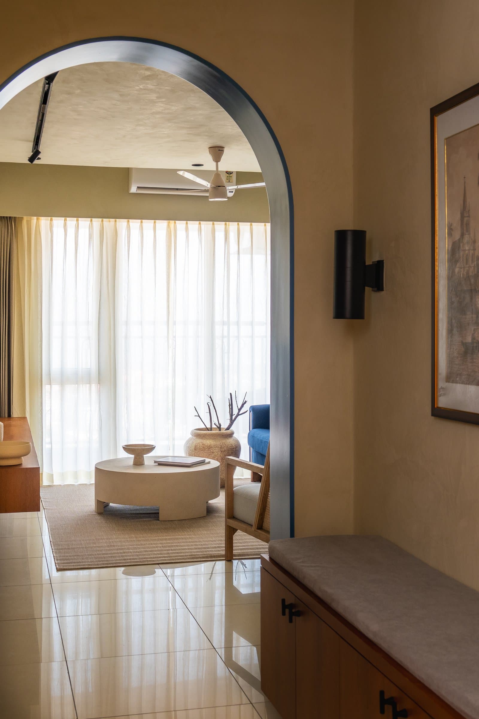The entry foyer: a timber storage bench and cylindrical black wall sconce prepare the arrival before the blue arch opens to the living room beyond