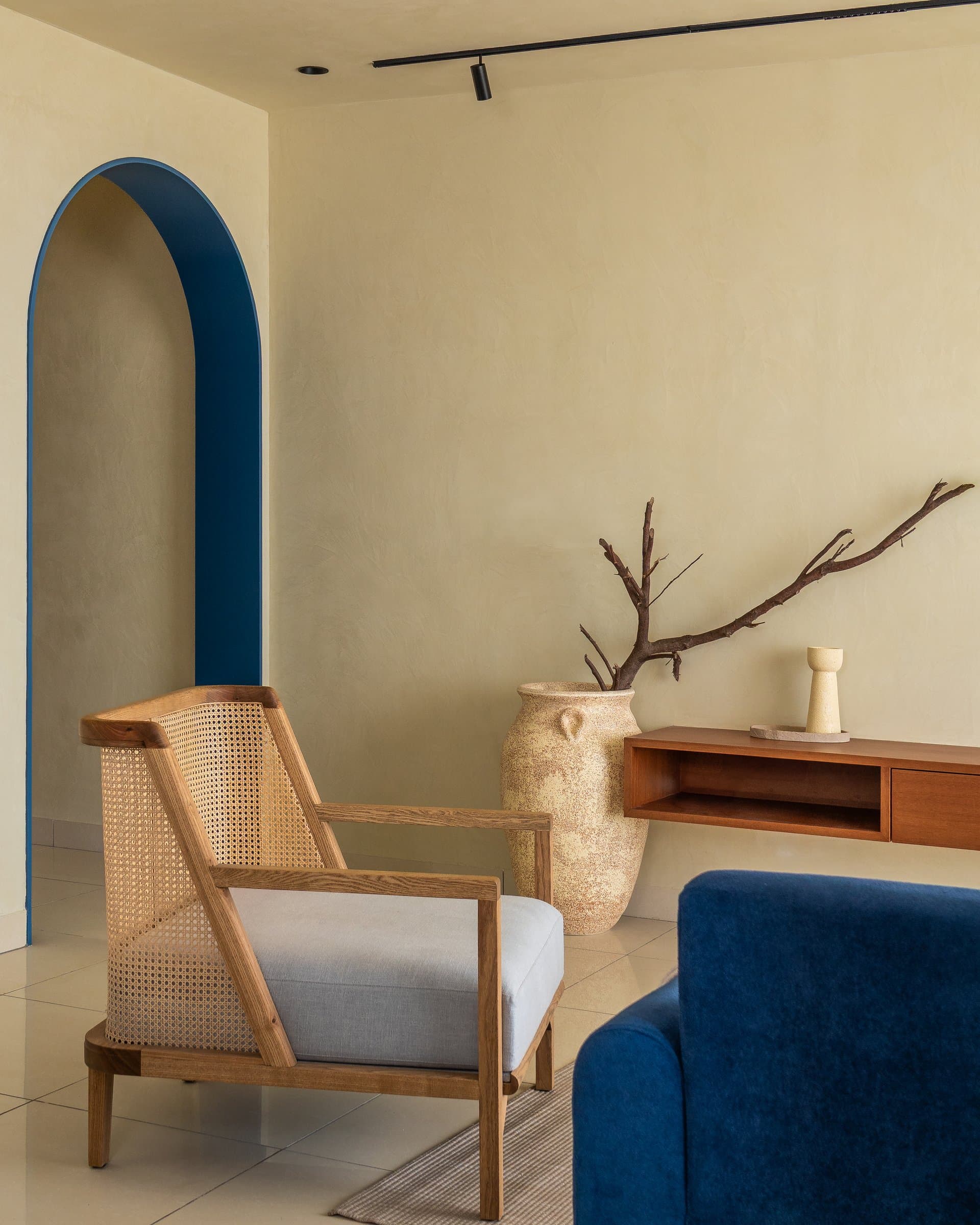 The blue-outlined arch frames the living zone's secondary corner, where a cane lounge chair and low timber console introduce the <em>wabi-sabi</em> note that runs through AOMI
