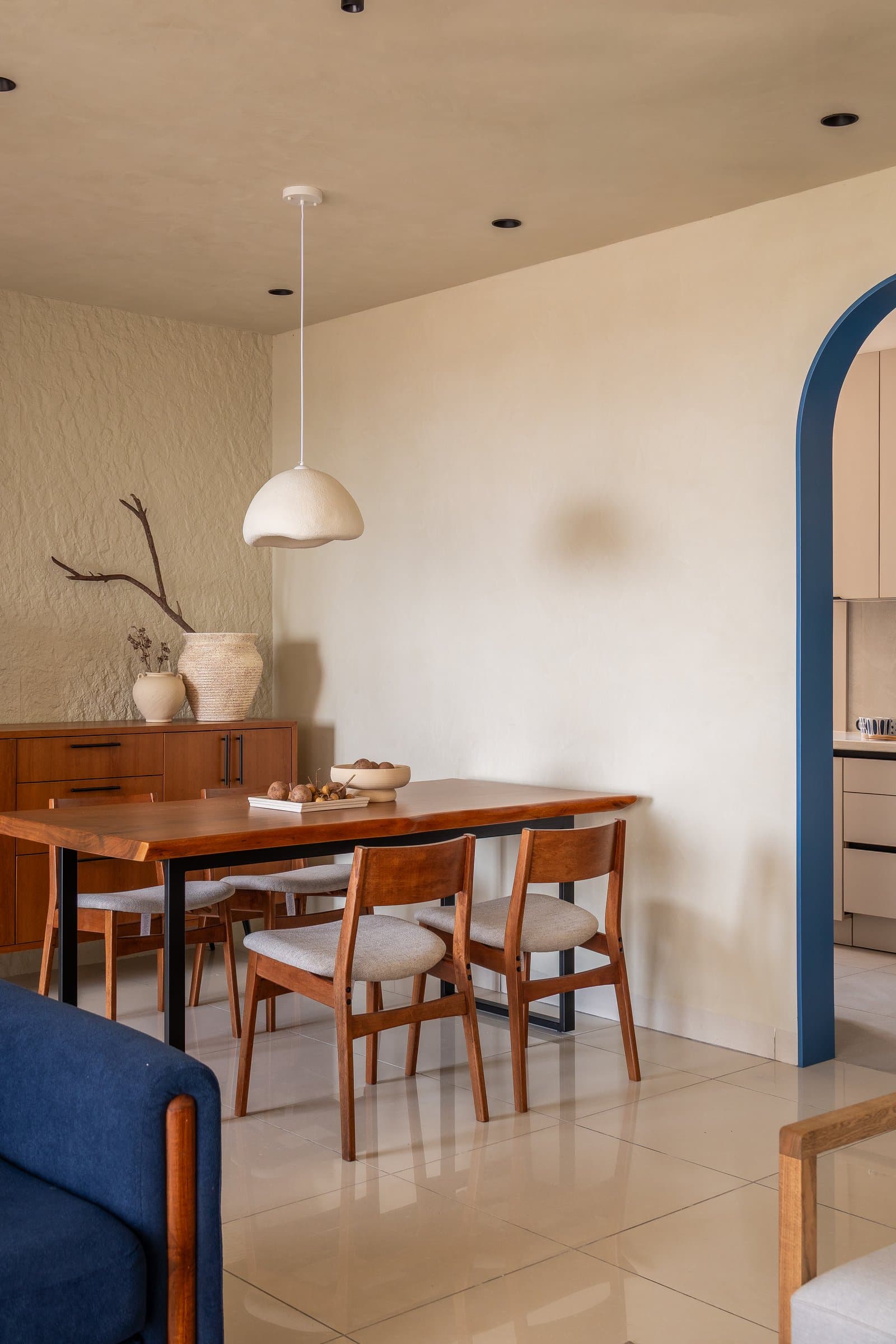 The dining area, where a live-edge timber table and a dome-shaped paper pendant share the frame with the blue arch and a rough-textured plaster feature wall