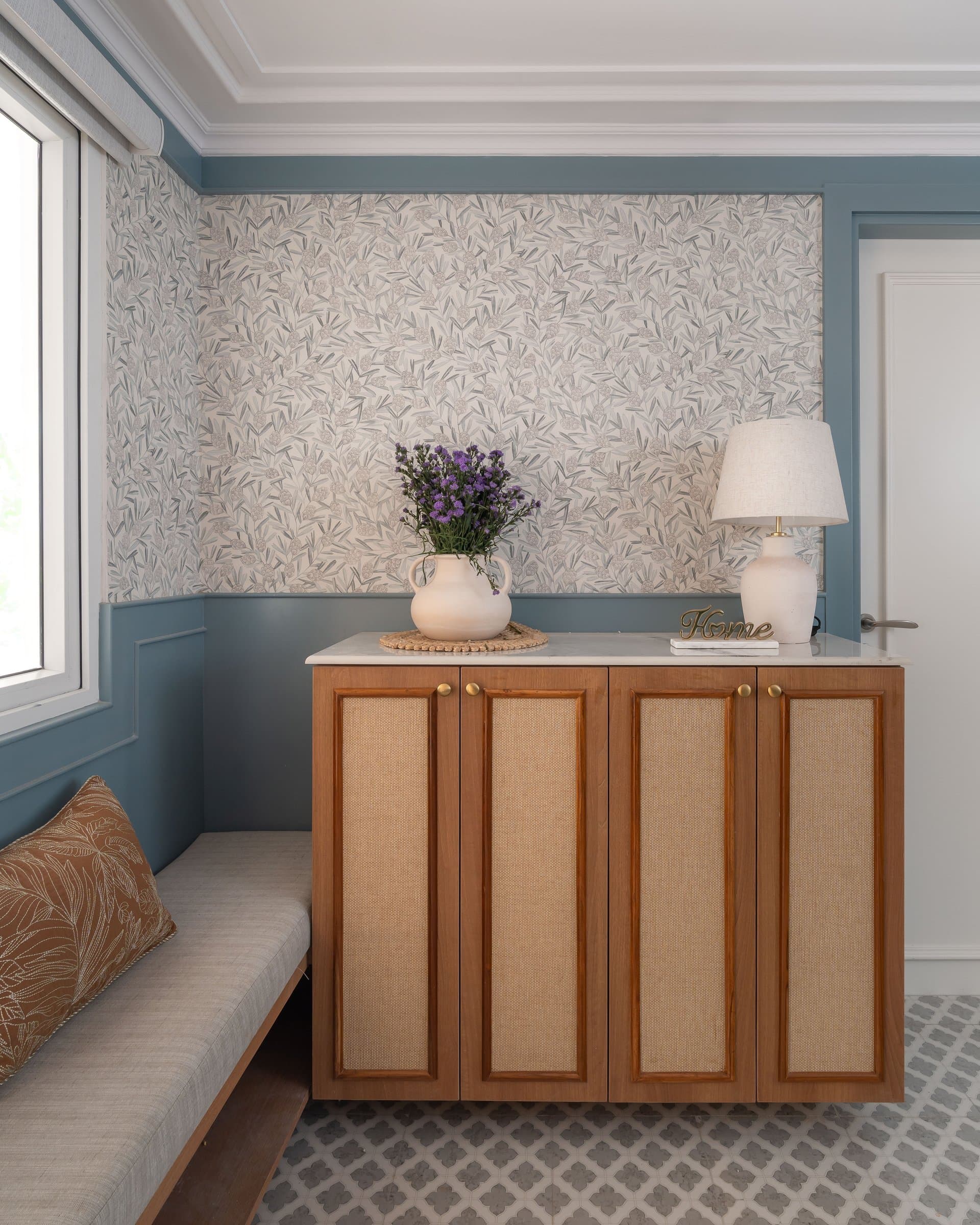The foyer: a cane-fronted console set against botanical wallpaper and dusty blue wainscoting establishes the home's appetite for pattern