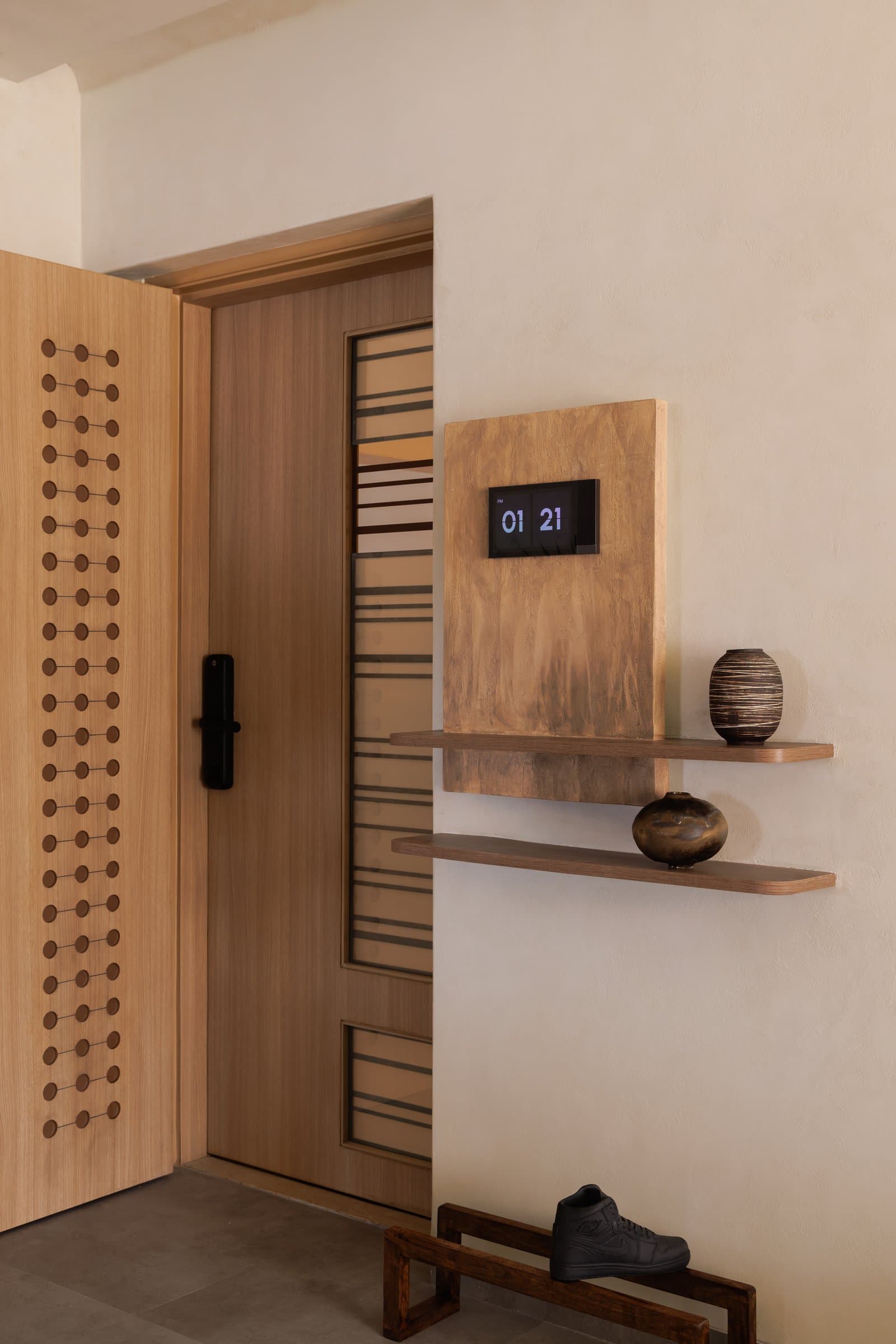 The entry, where a perforated wooden door and a wood-mounted flip clock establish the home's tactile, time-slowed grammar