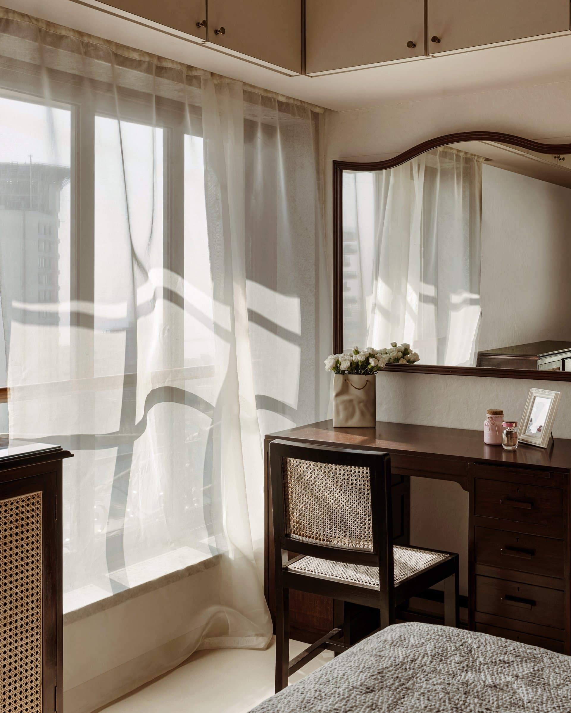 A dressing corner where utility becomes stillness: a dark wood desk with an arched mirror, a cane-backed chair, and late light through sheers