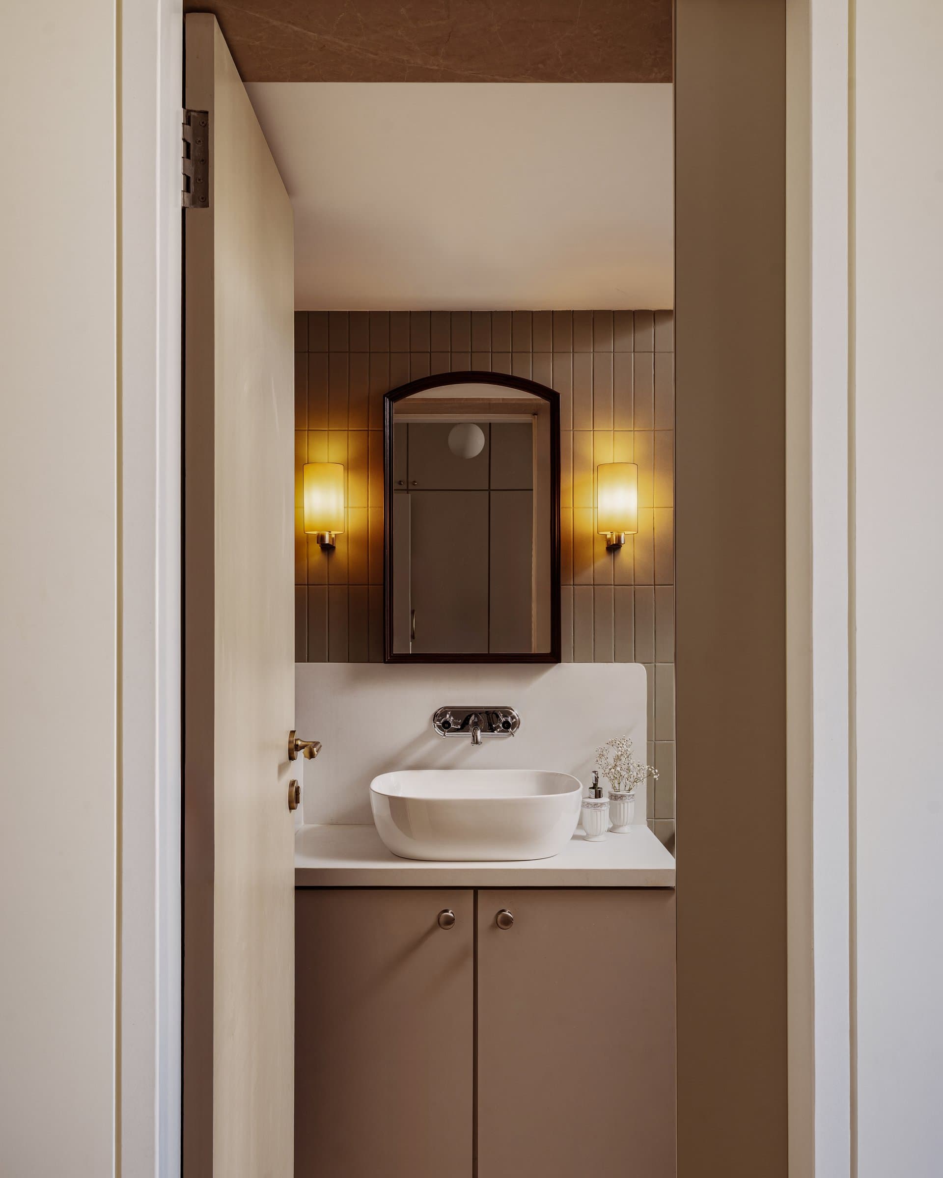 The bathroom, where olive-toned vertical tiles, a soft mauve-grey vanity, and an arched mirror close the project on a precise note