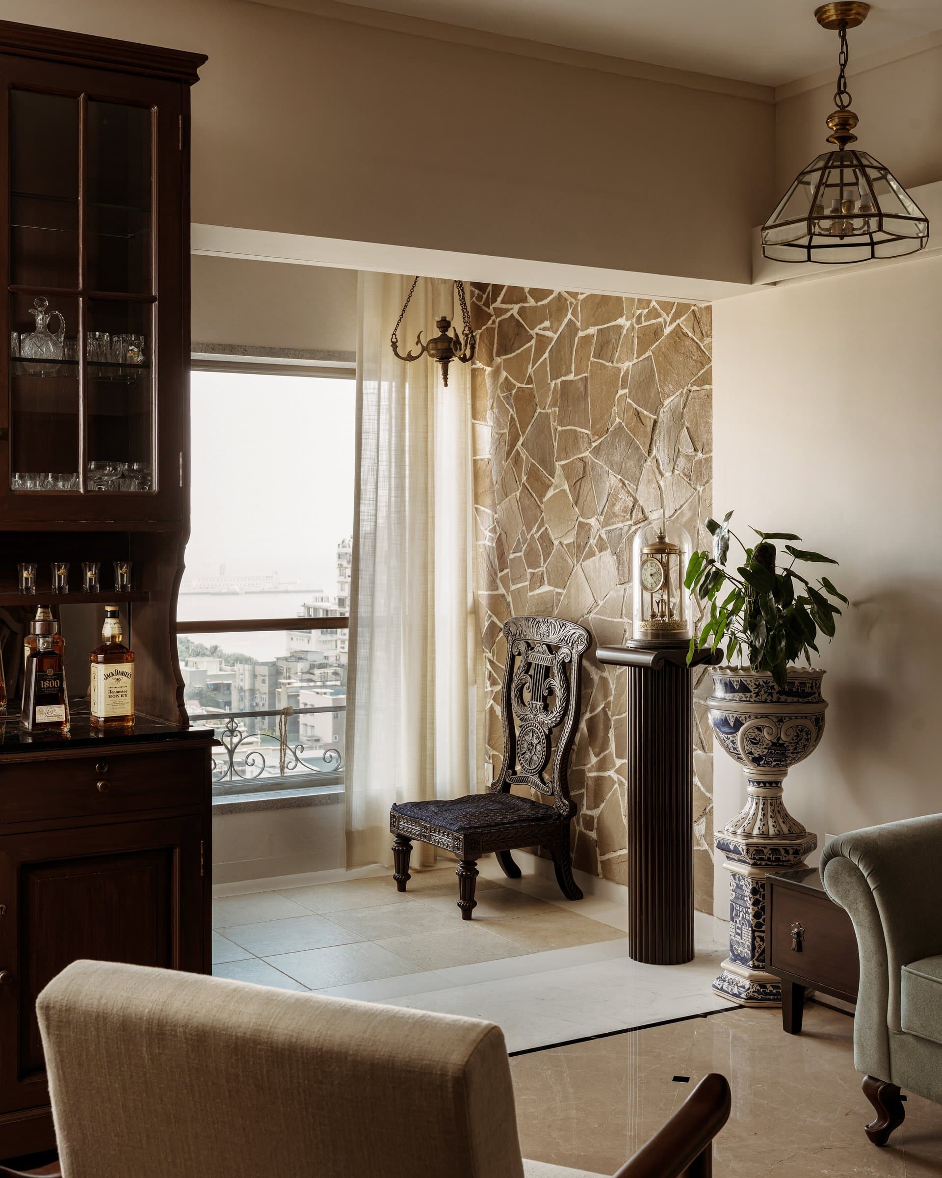 A corner of the living room: a wall of hand-laid stone meets a heritage carved chair and a fluted pedestal, the city skyline filtering through sheer curtains