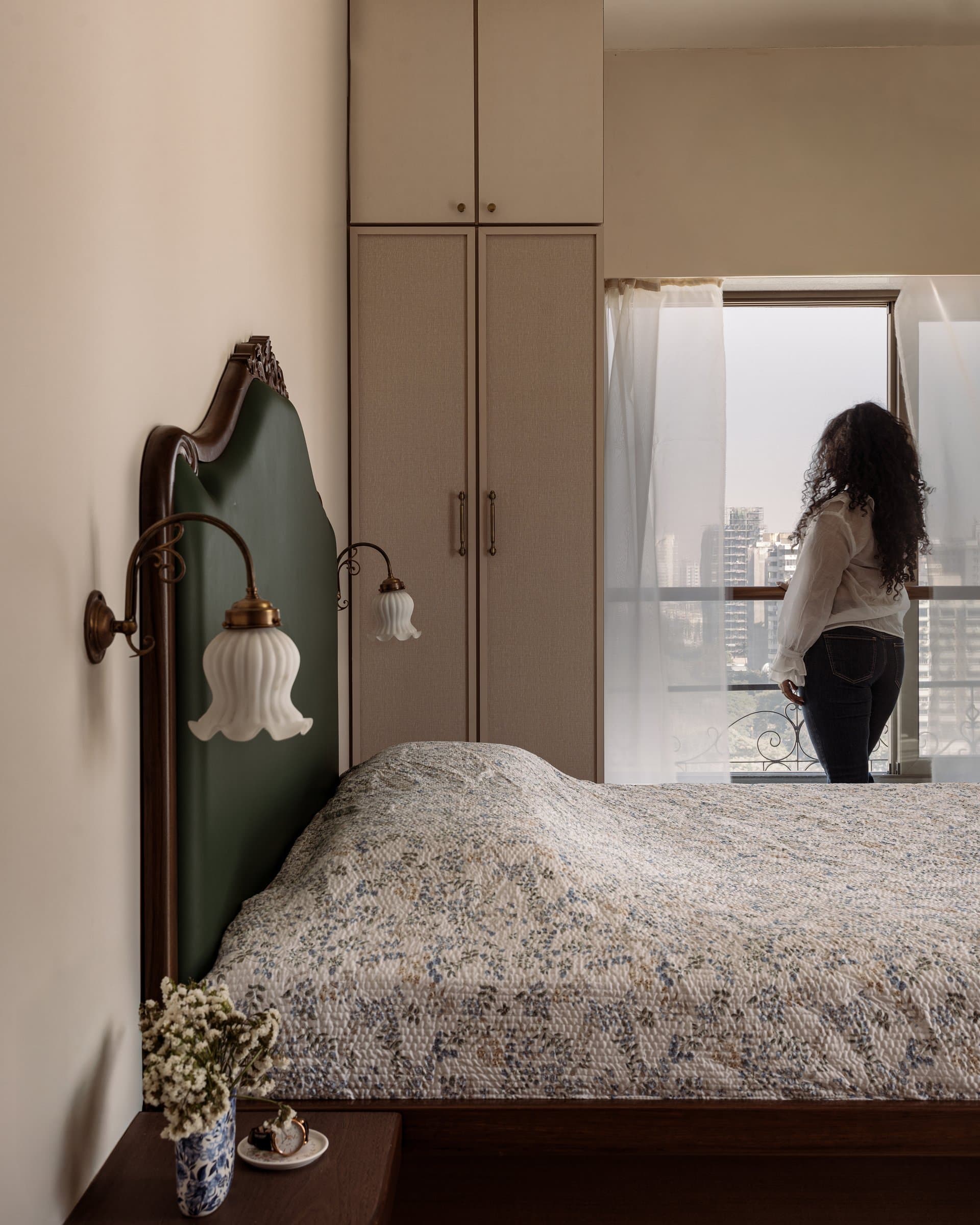 The bedroom, anchored by a forest-green upholstered headboard, brass tulip sconces, and a fabric-fronted wardrobe in oat tones