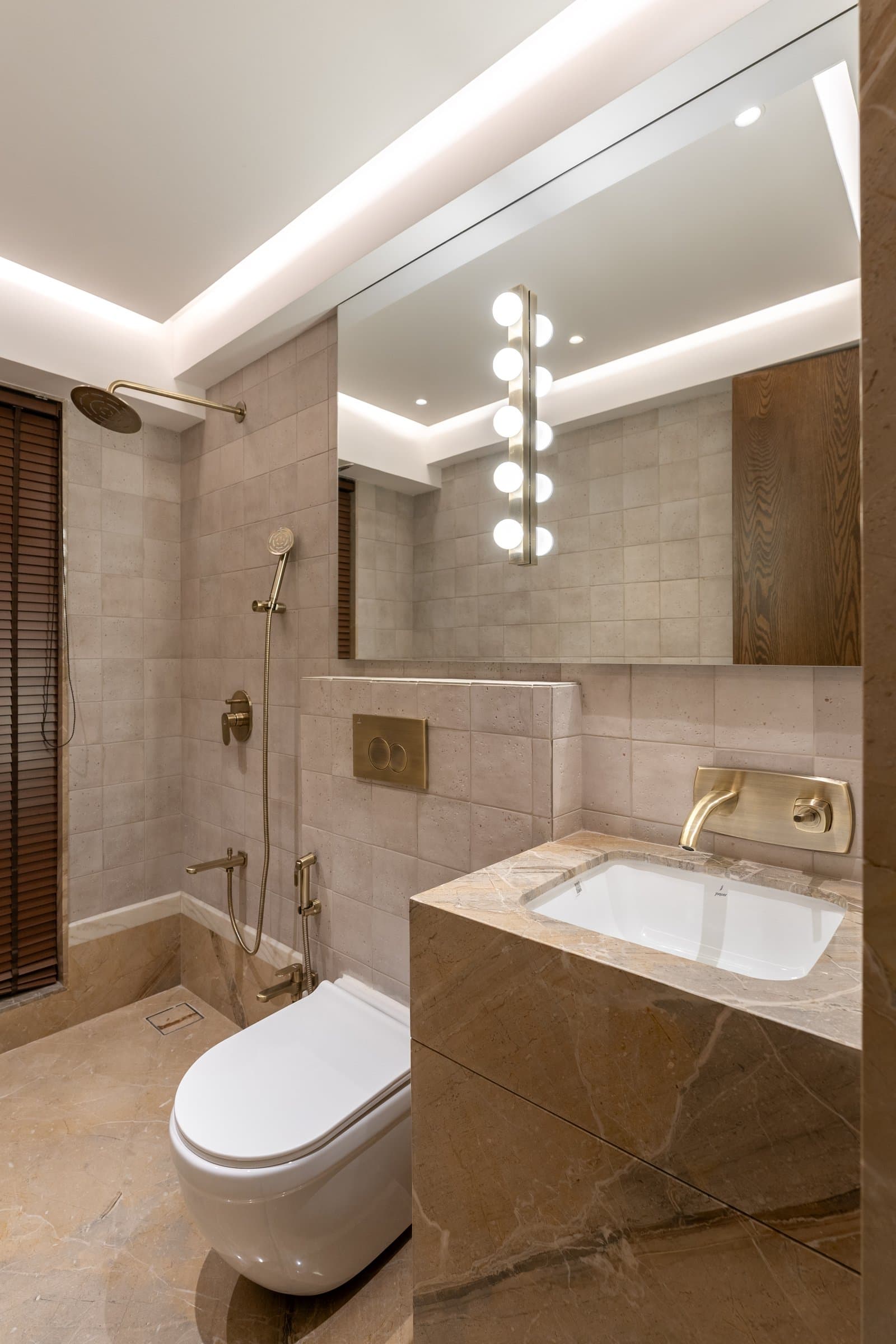 A bathroom in honey-toned stone, with antique brass fittings and a louvred wooden screen at the window