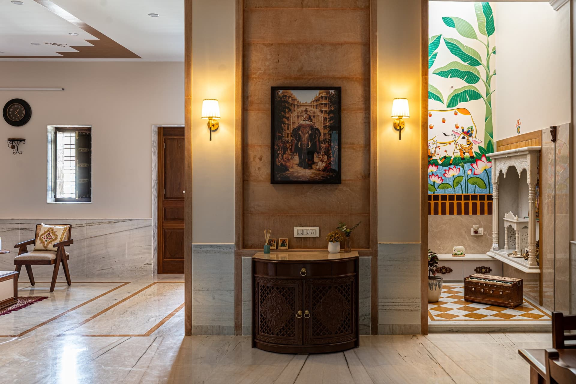 The transition zone between living and pooja room, sandstone pier and dark-wood console on one side, Pichwai mural and marble shrine on the other