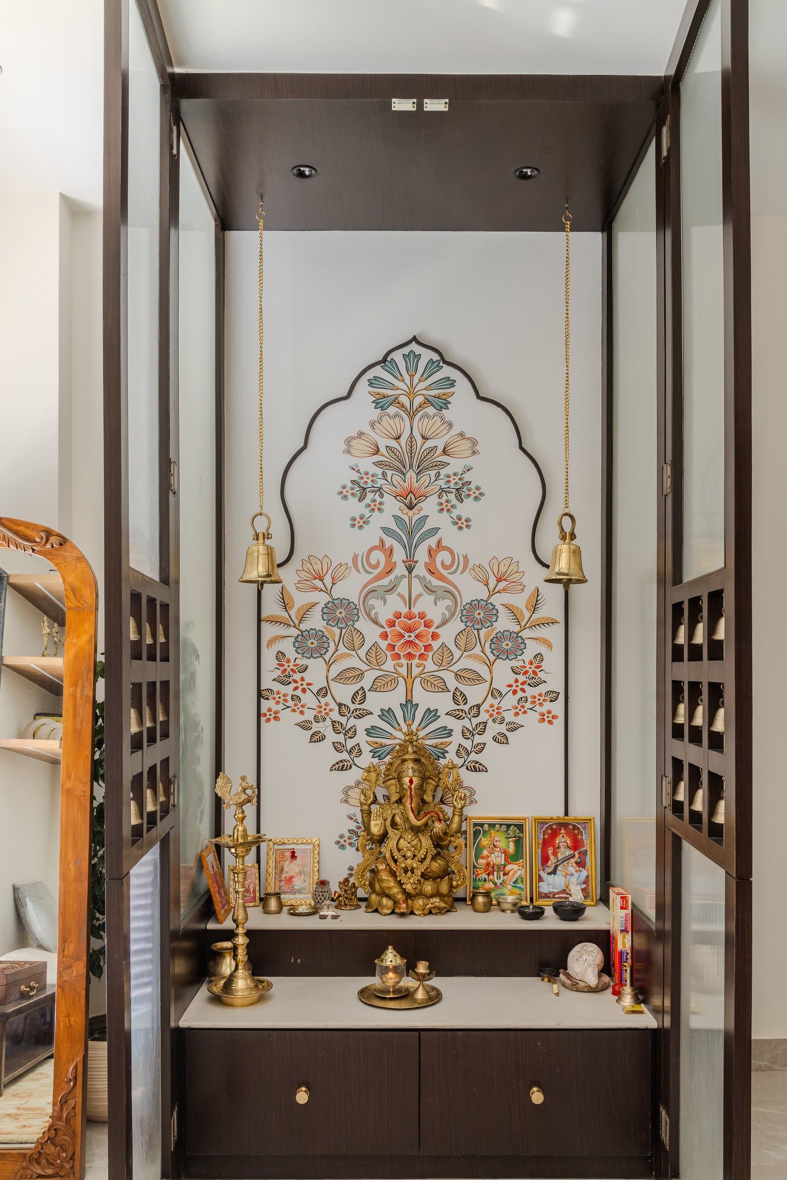 The mandir, framed in dark wood and fluted glass, with a floral wallpaper rising within an arched outline behind the brass deities