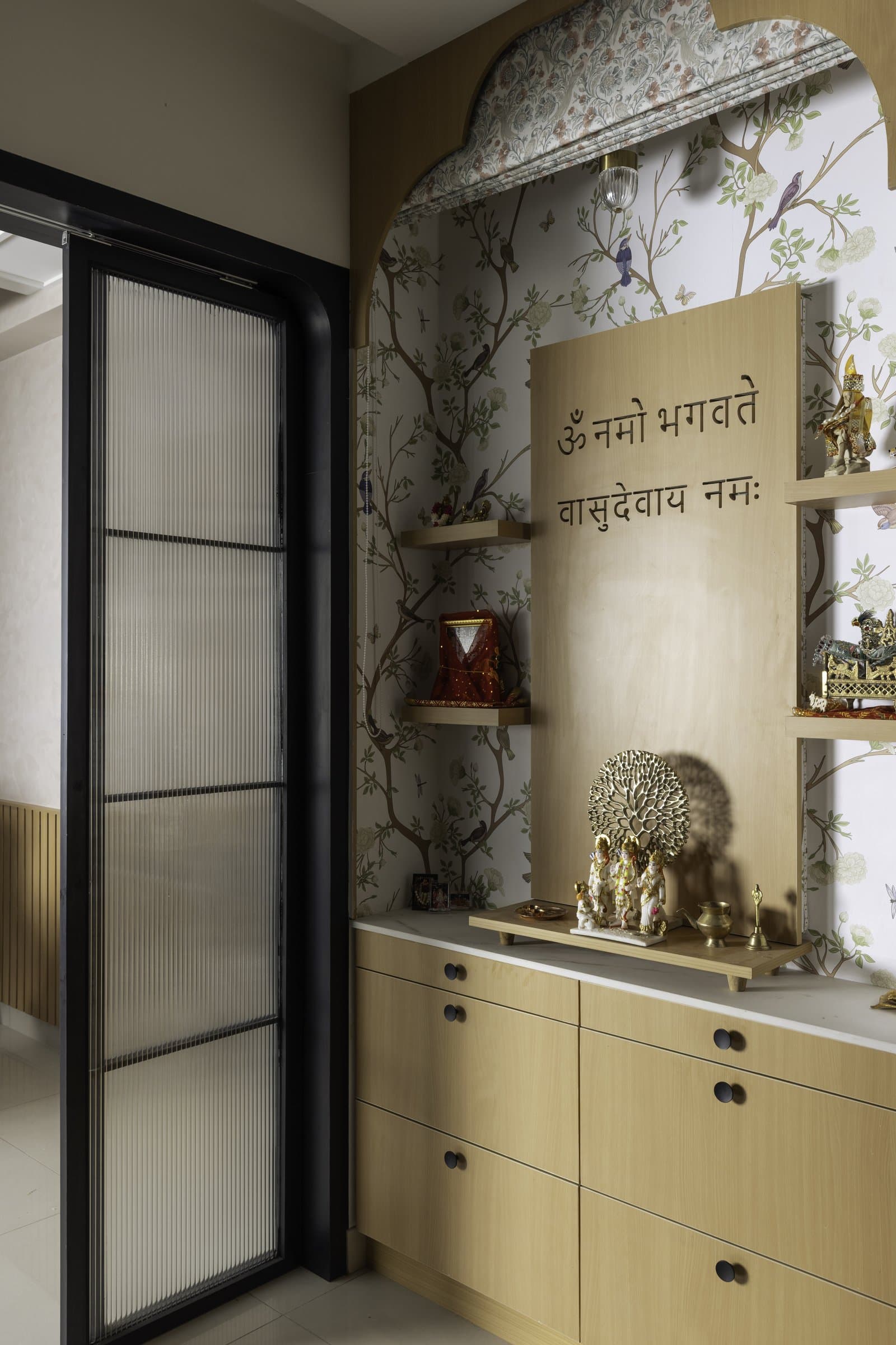 The puja room behind a fluted glass sliding door, its chinoiserie wallpaper and Sanskrit invocation marking the home's quietest pause