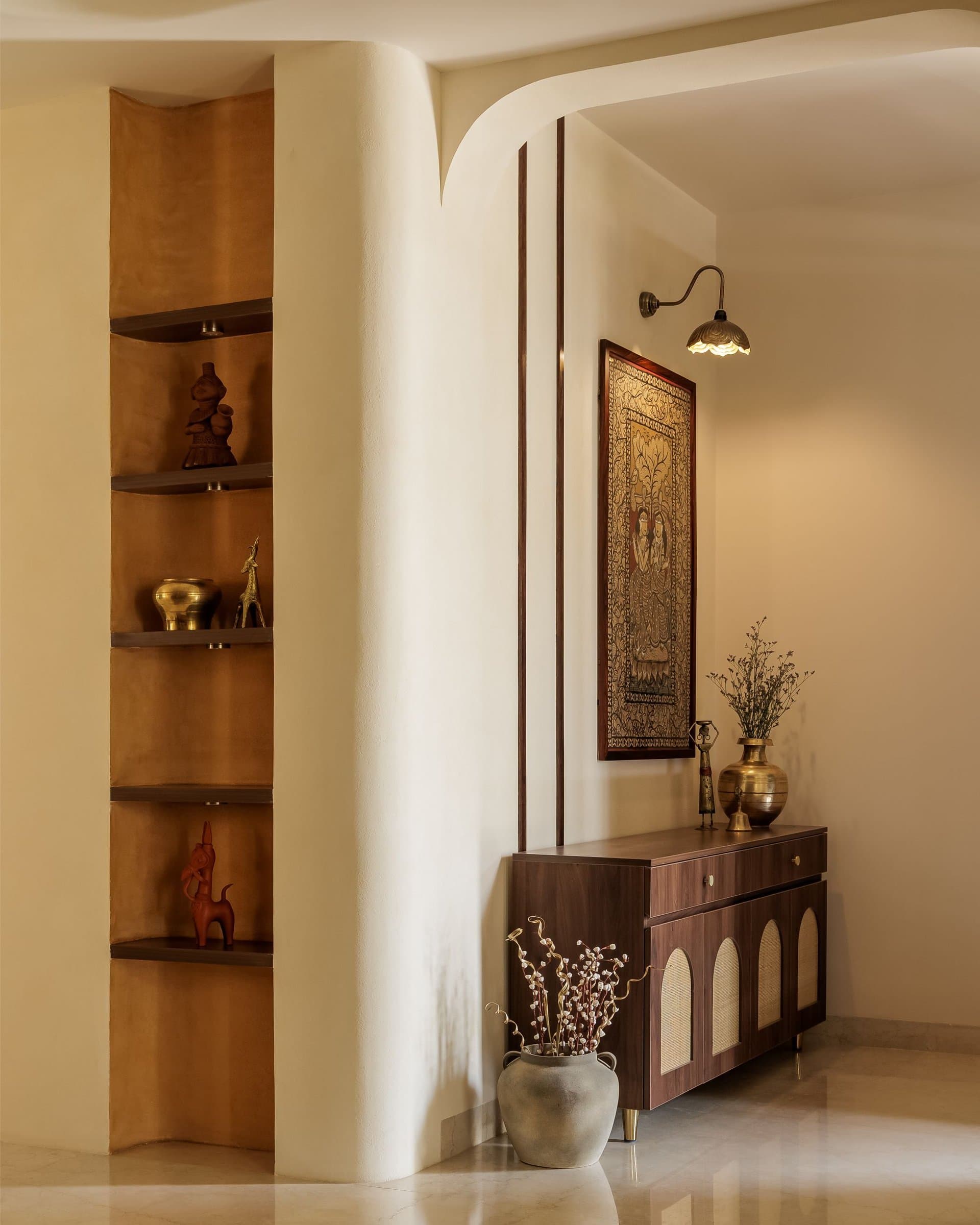 The entry, where a rounded ochre pier of recessed niches holds folk objects opposite a cane-fronted console and framed Indian artwork