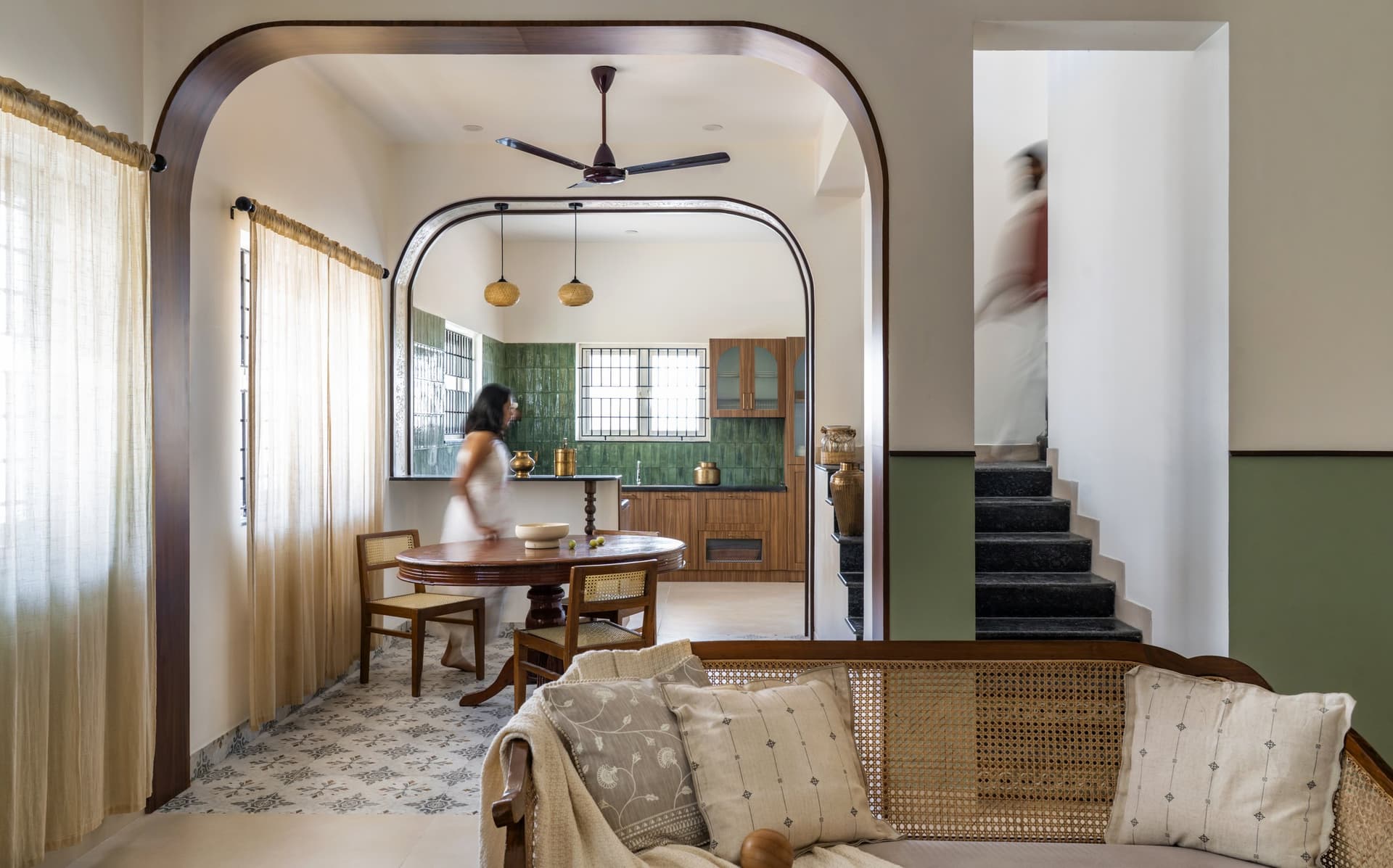 The opening view, where a rounded wooden arch frames the dining and kitchen beyond a caned settee in the living room