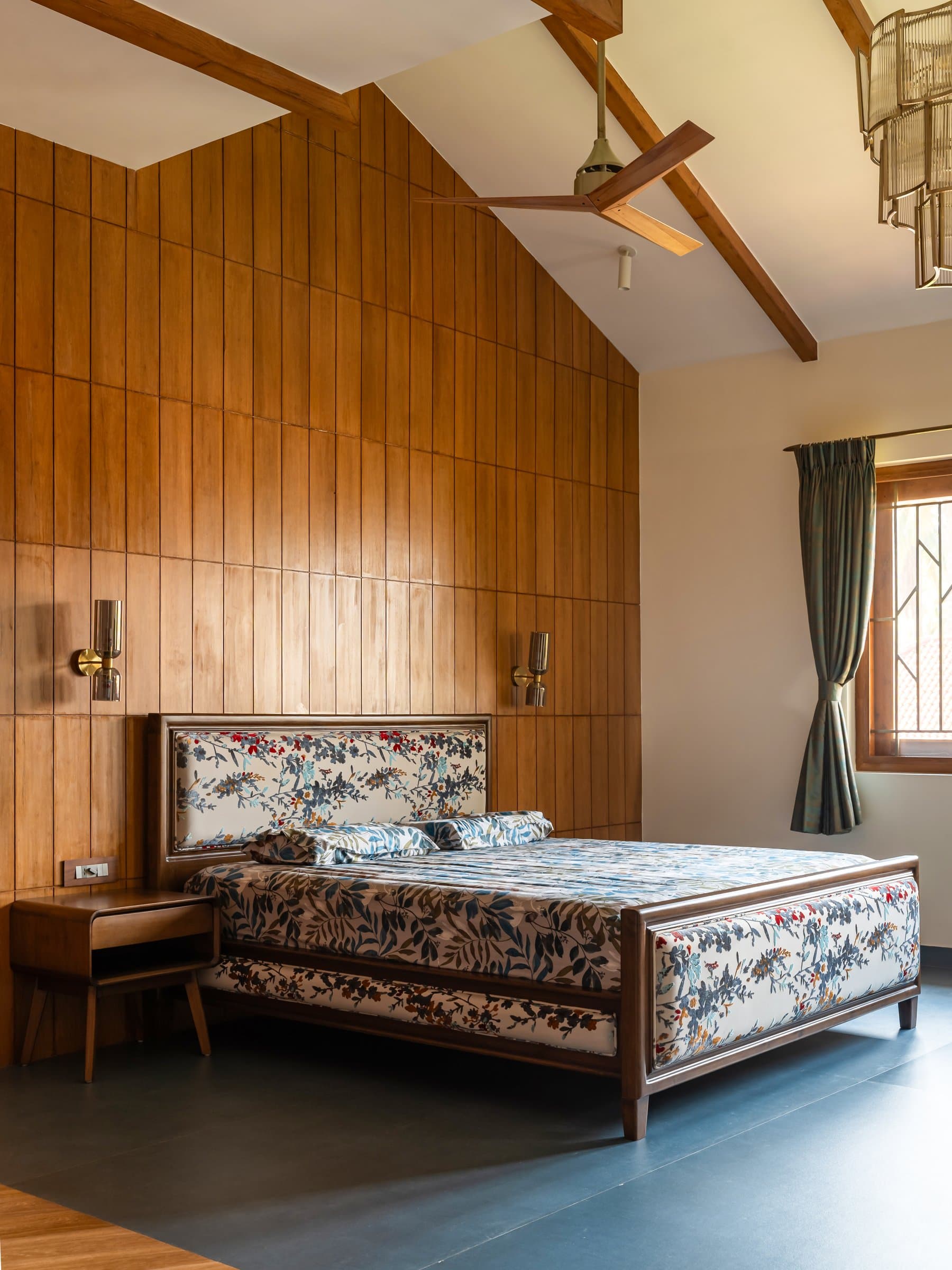 An adjacent bedroom under the same pitched roof, lit by paired brass sconces on a full-height timber wall