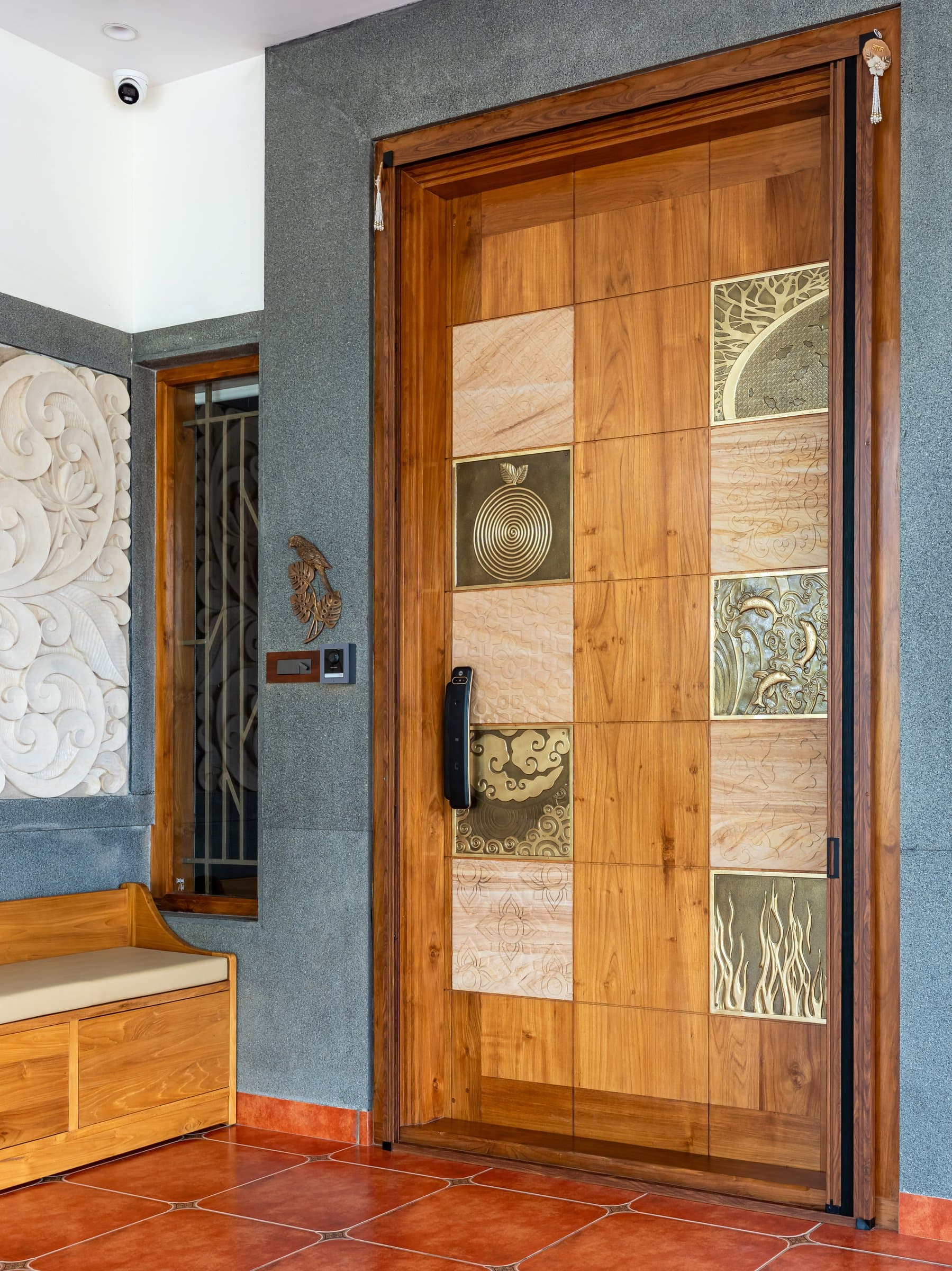The main door: a teak grid inset with brass motif tiles, framed by a carved stone bench and lotus relief