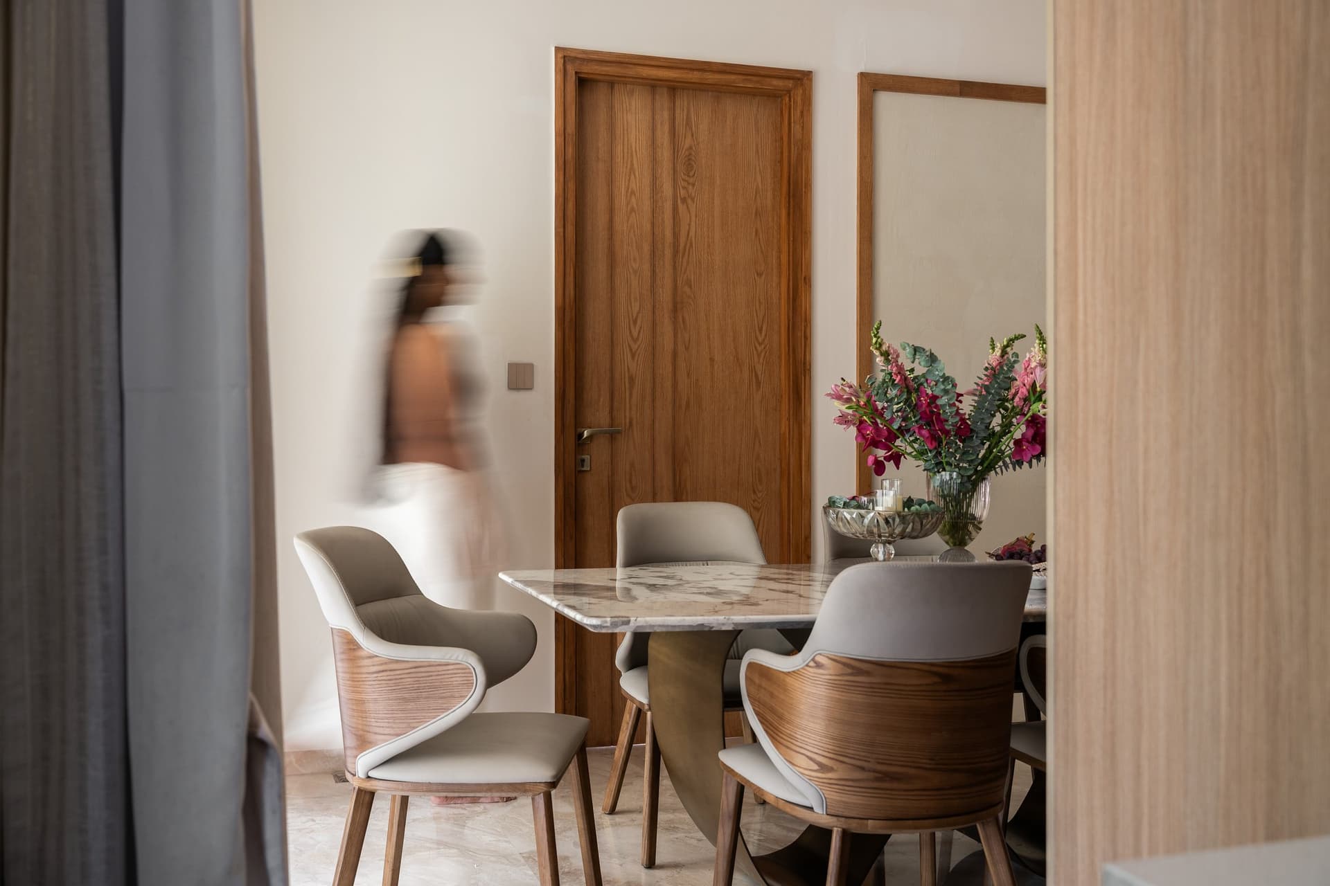 The dining area, glimpsed through a curtain edge, where taupe leather chairs surround a marble-topped table