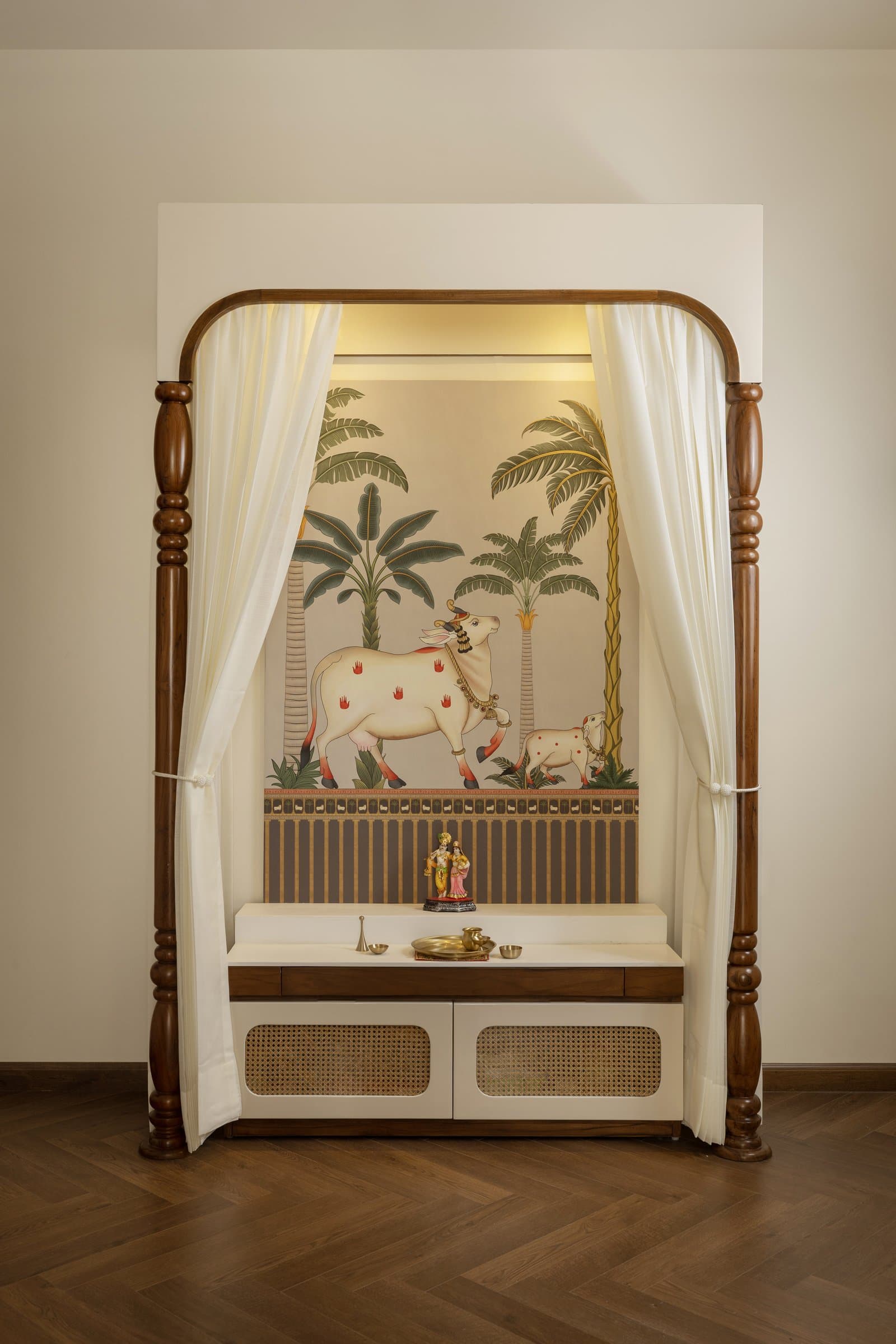 The pooja niche, framed by turned wooden columns and white sheers, with a Pichwai-inspired mural behind a cane-fronted counter