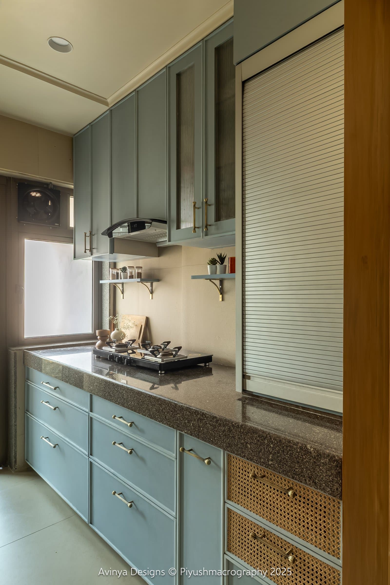 The galley kitchen, where sage-blue panelled cabinetry meets a dark speckled stone counter and cane-fronted lower drawers