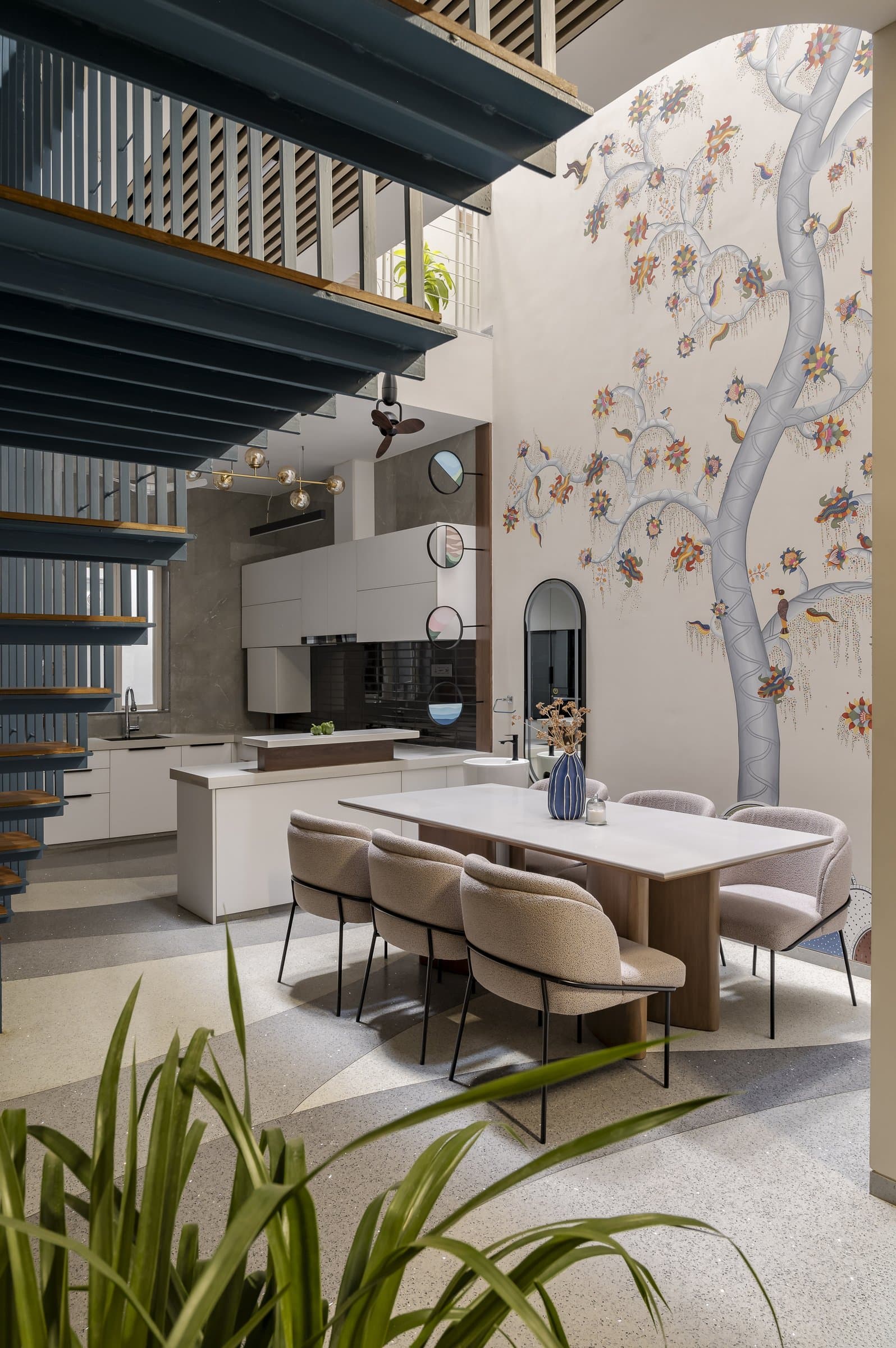 The dining area beneath the triple-height void, where a Madhubani-style mural climbs alongside the exposed blue steel structure