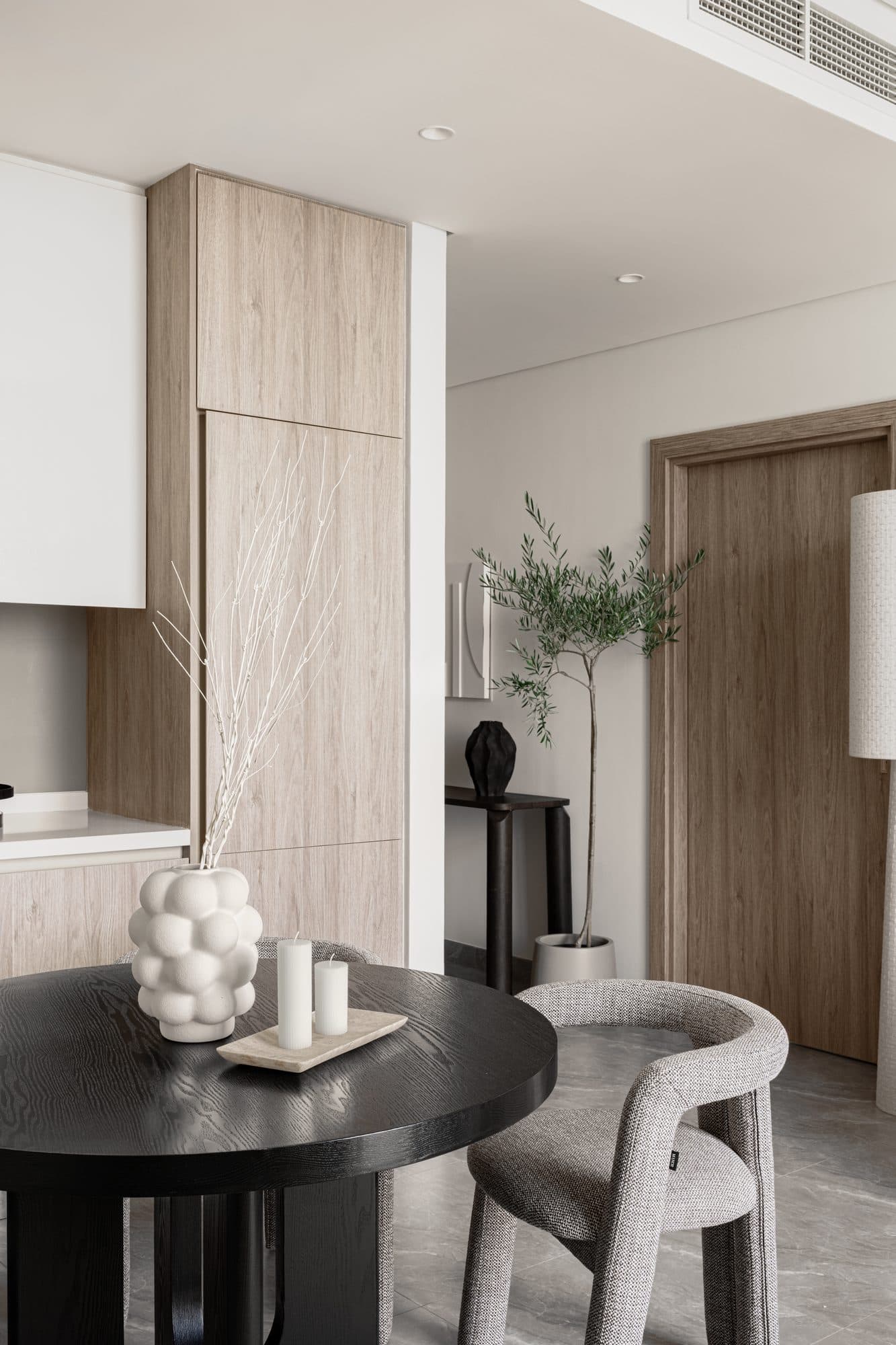 Dining as still life — a black-oak round table, a sculptural bud vase, and warm wood cabinetry against grey stone floor.