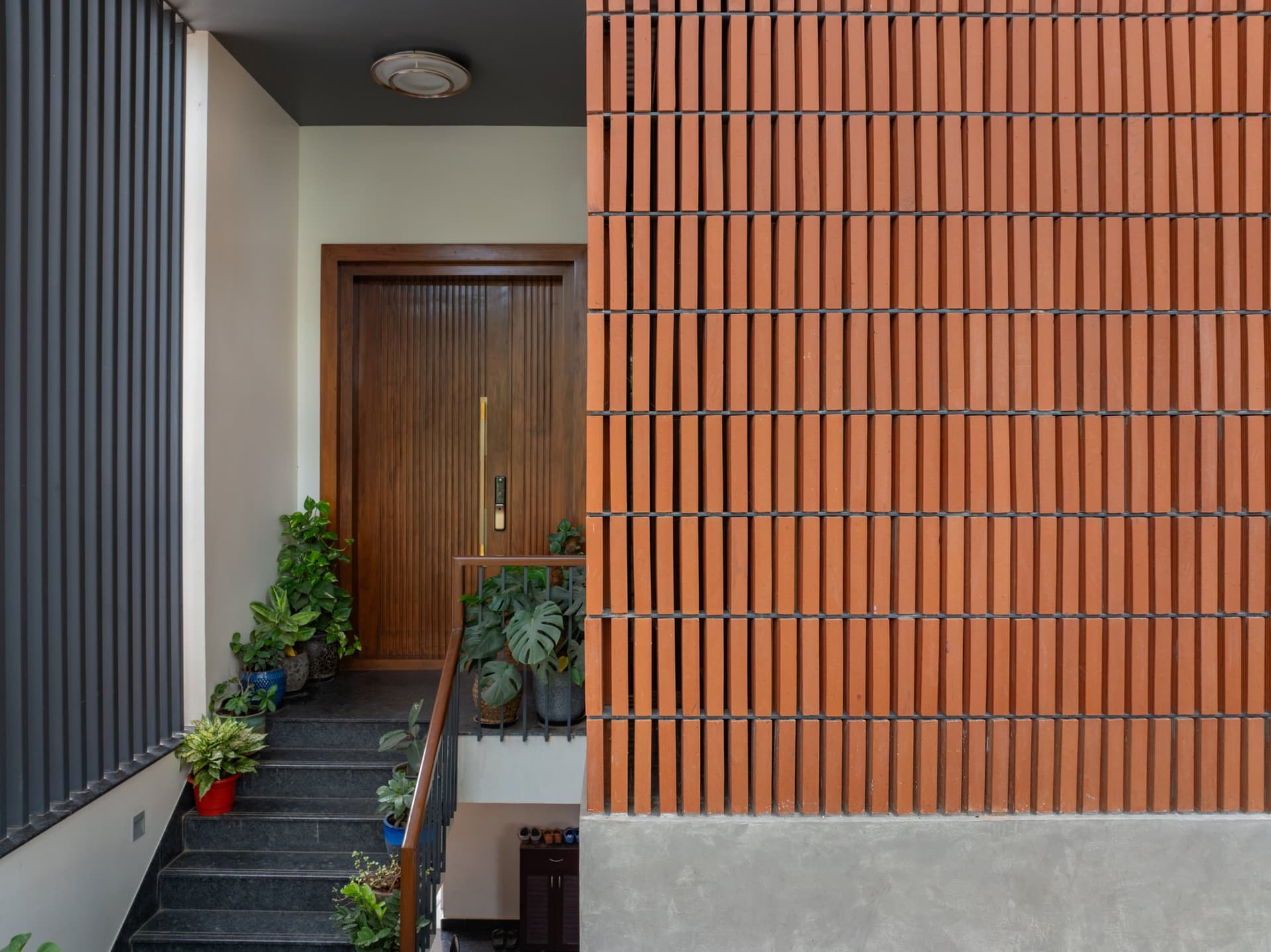 The entry threshold, where a perforated terracotta brick wall meets a slim aluminium fin screen along the granite ascent to the front door