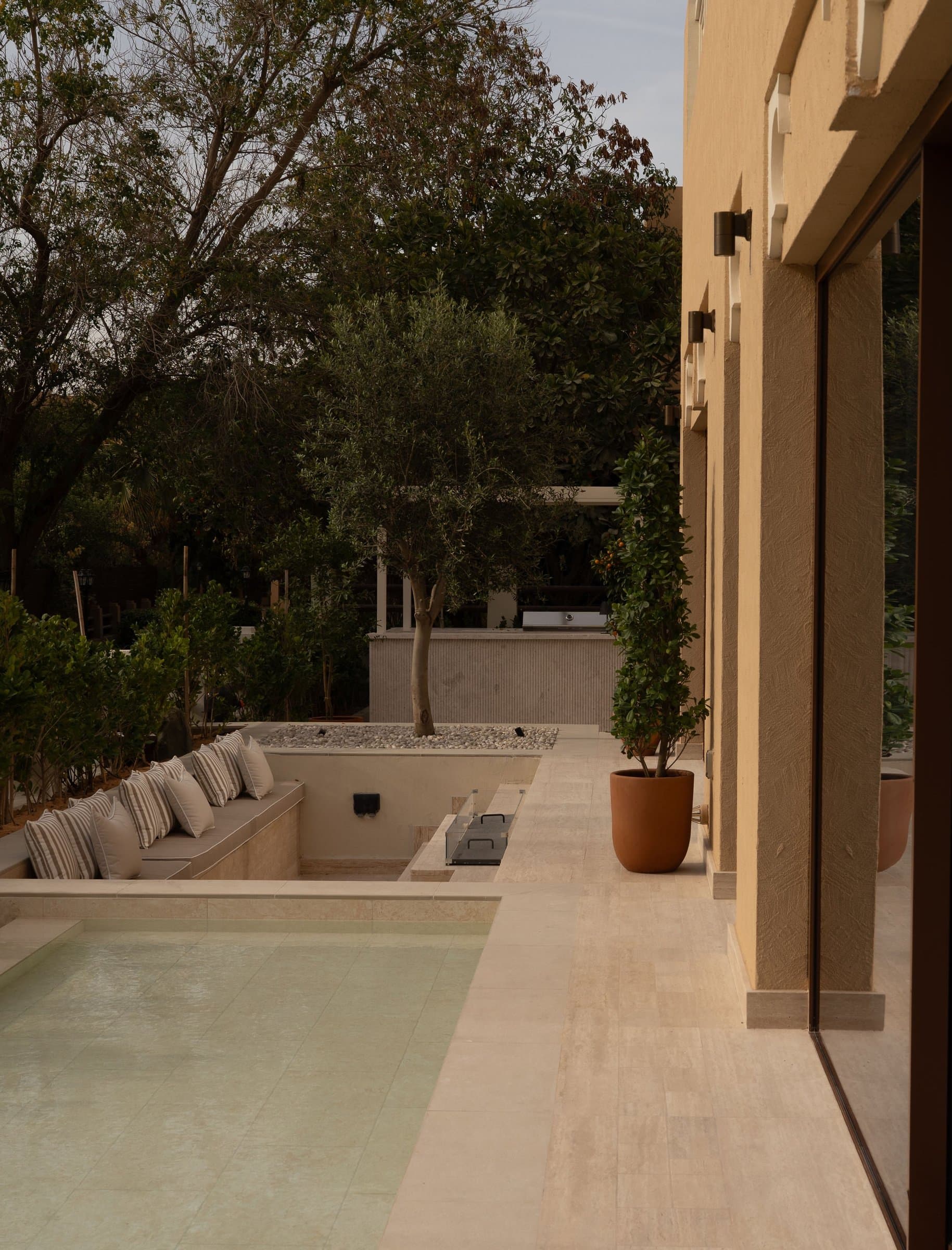 The terrace runs alongside the villa's plastered facade toward a sunken pool sheltered by an olive tree.