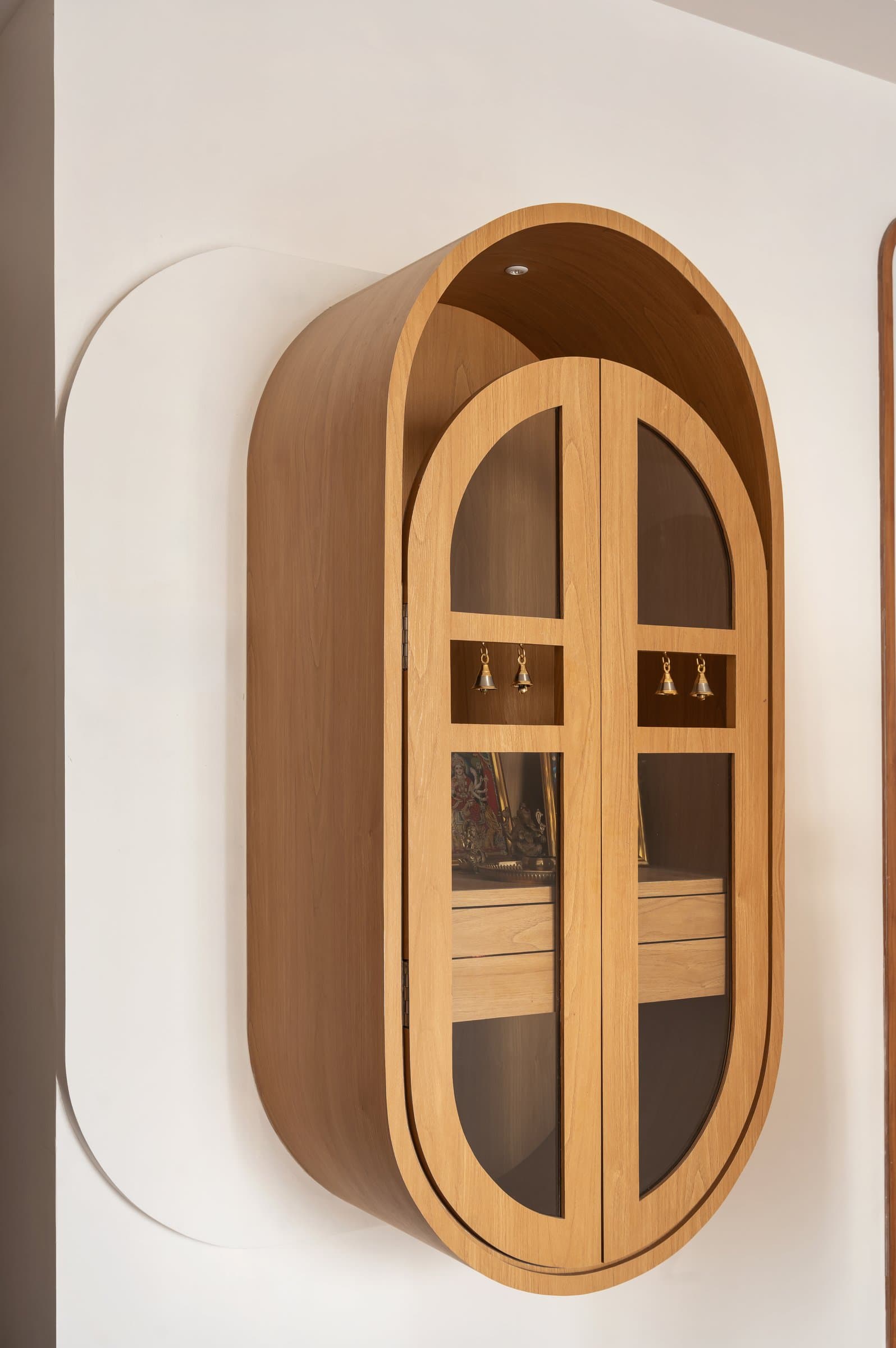 The freestanding wooden puja unit, a capsule silhouette with arched glass doors against a scored wall profile