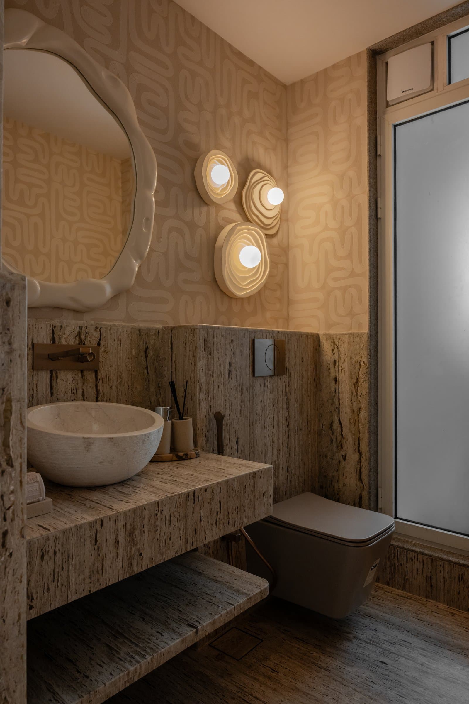 The powder room, where travertine, patterned wallpaper and organic ceramic sconces compose a sculptural interlude