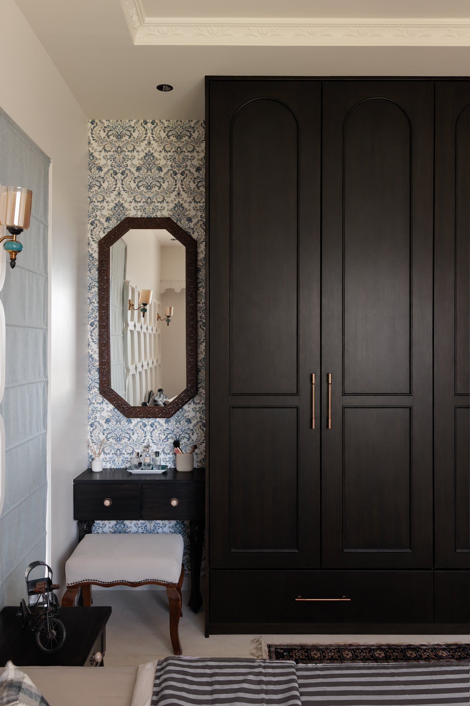 The dressing nook, where a dark wooden vanity and tall wardrobe share a wall with a patterned wallpaper in blue and ivory