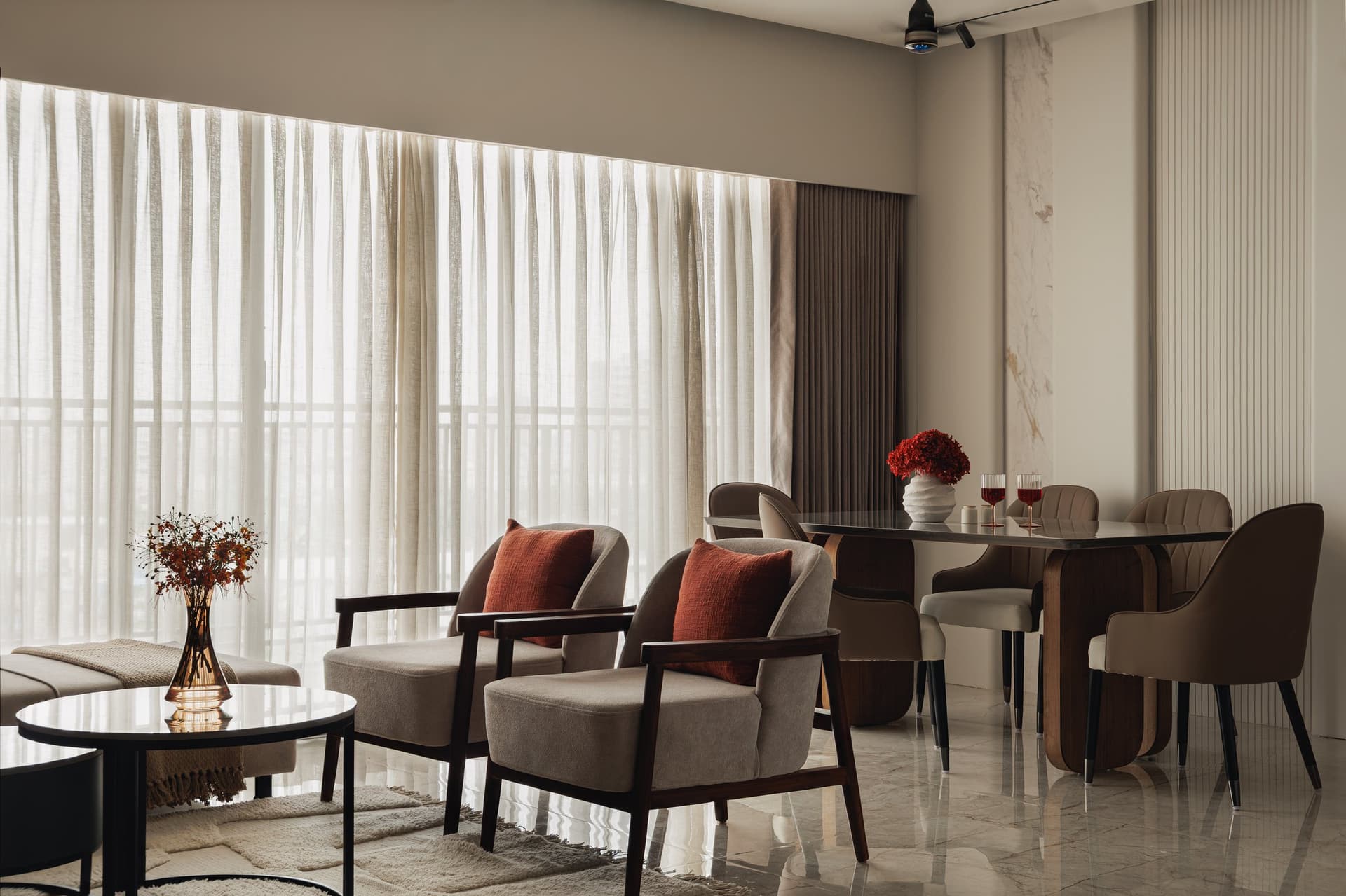 A wider view connecting the dining area with the adjacent lounge seating, where timber-framed armchairs sit beside sheer-curtained windows