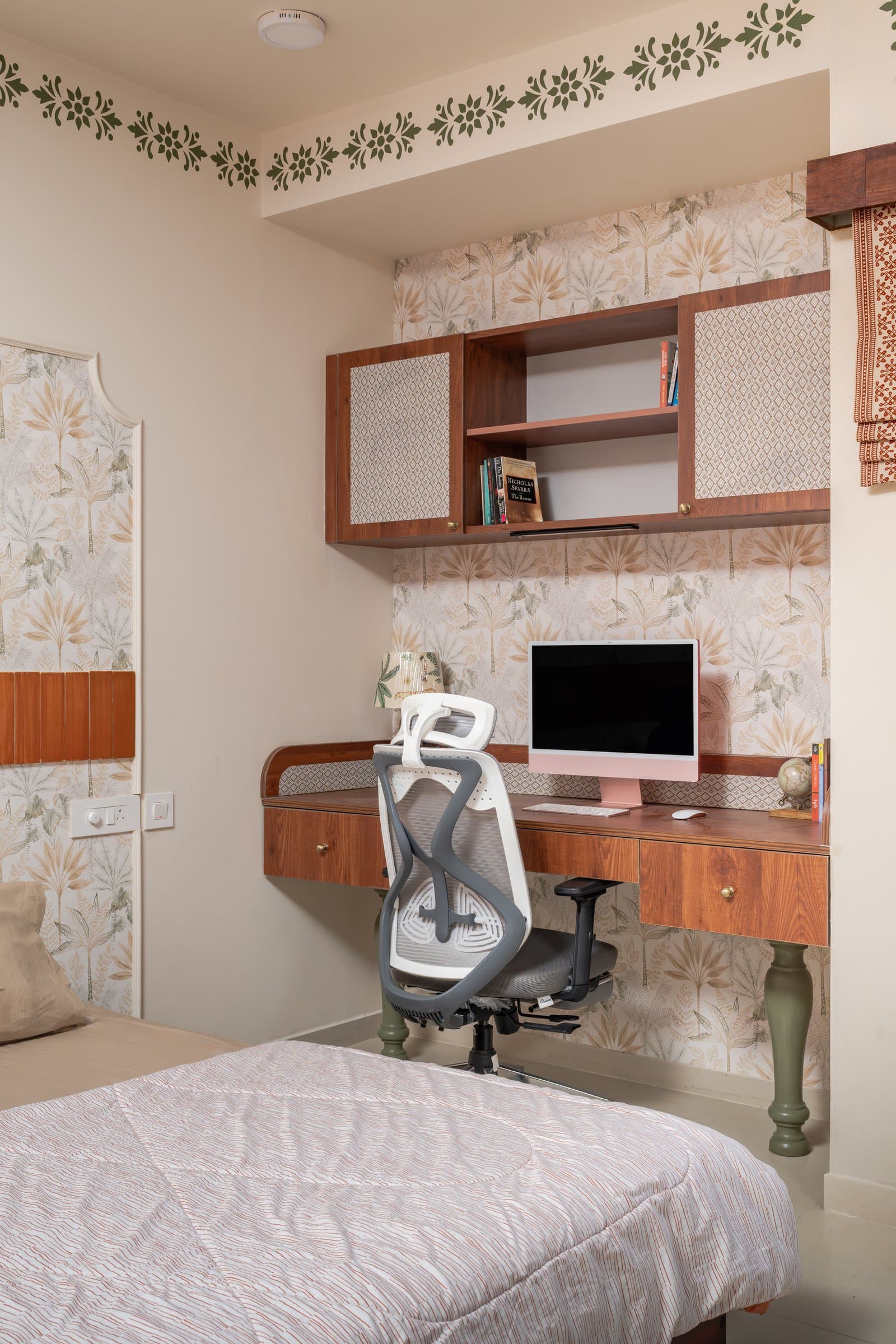 A compact study niche within the bedroom, with a wooden-topped desk and turned sage-green legs set against patterned wallpaper