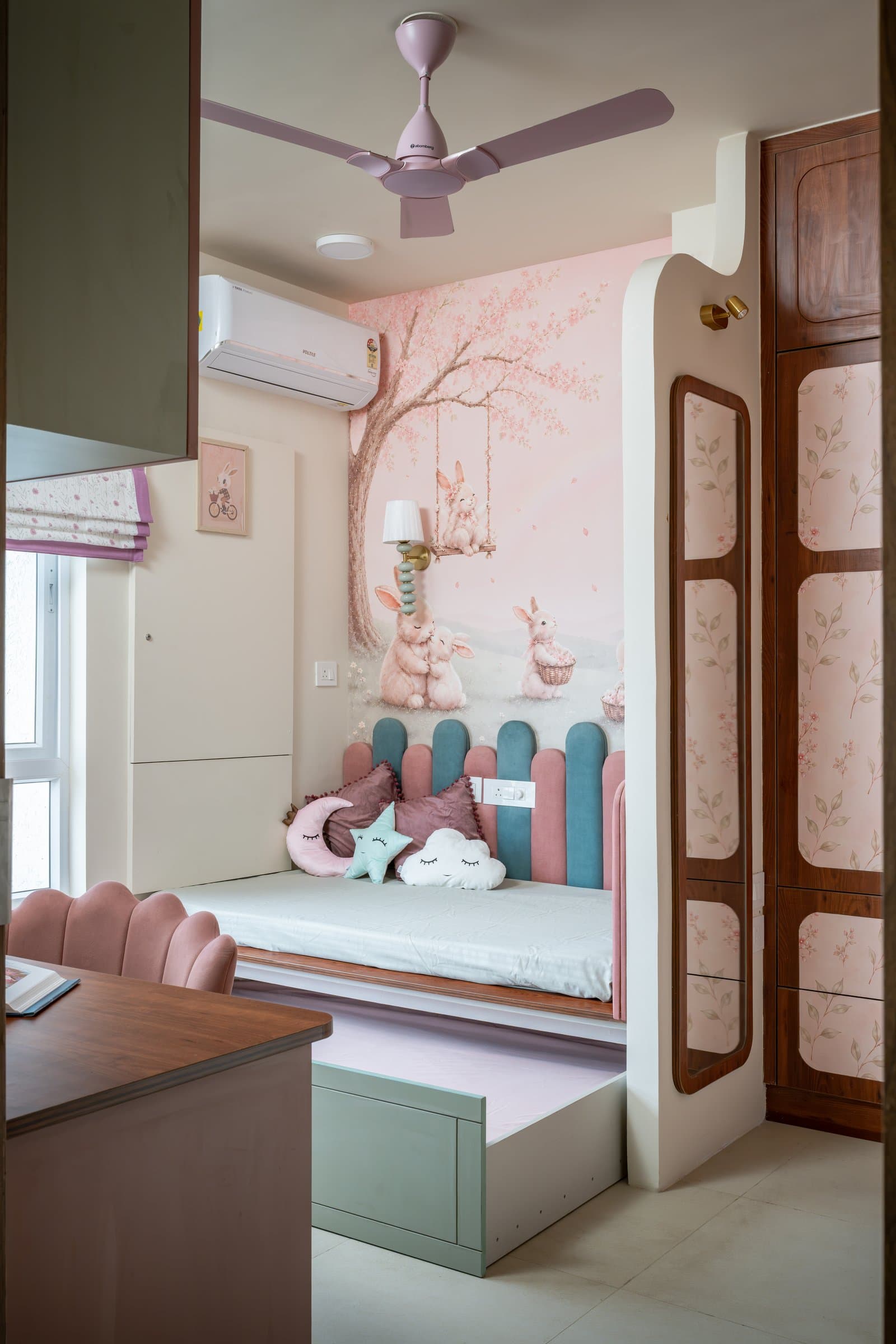 The children's room, with a patterned wallpaper above a window seat and a headboard made of alternating blush and teal upholstered pickets
