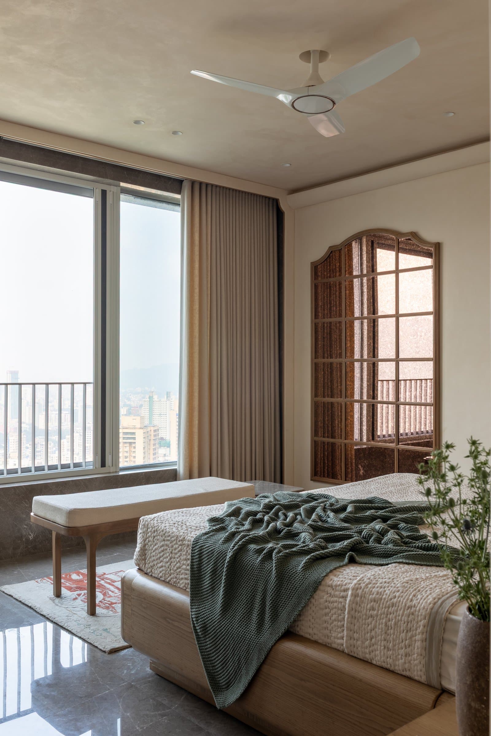 At the foot of the bed, a wooden-framed bench sits beside full-height glazing that opens the room to the city