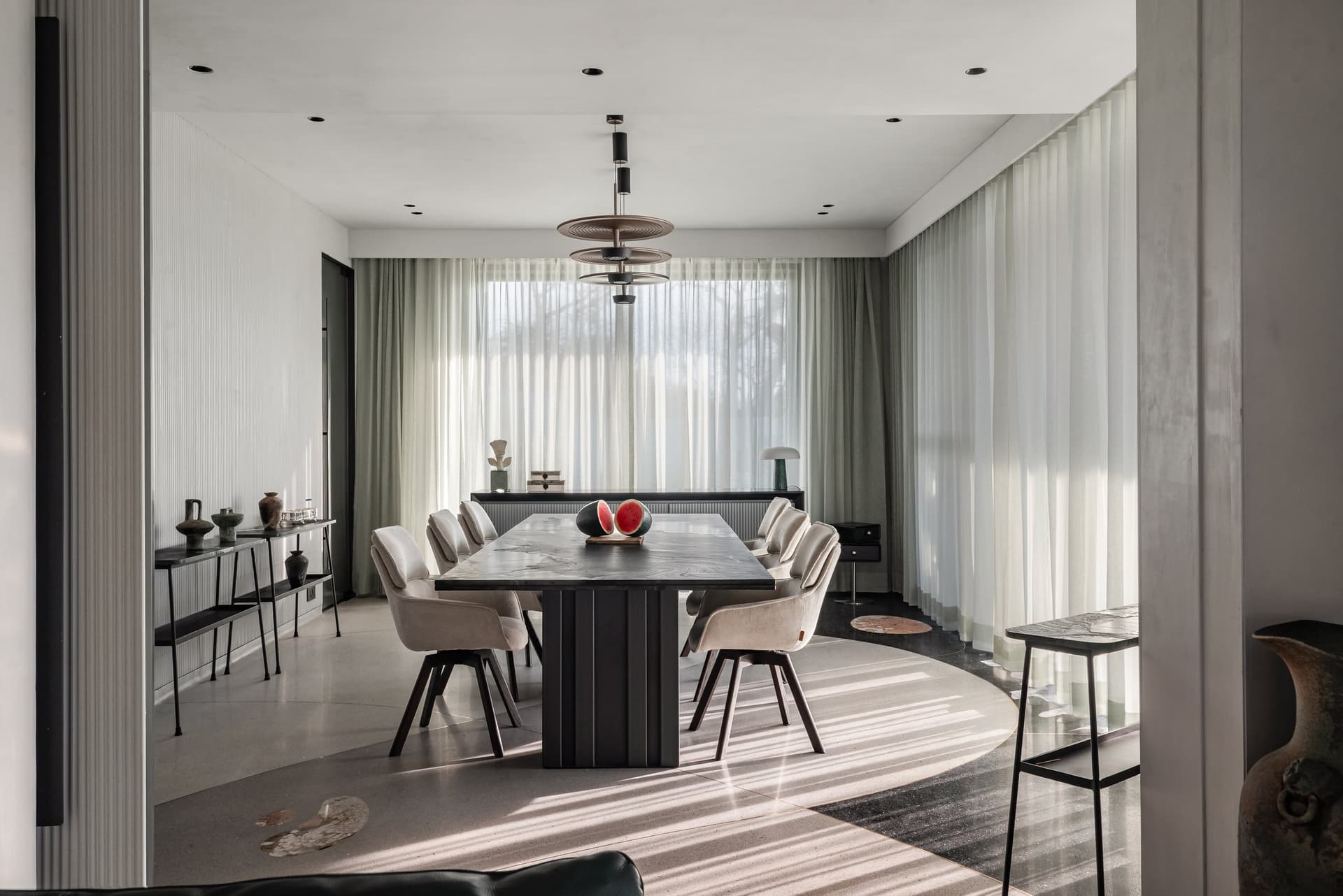 The dining room seen straight on: long table, tiered pendant, and afternoon light striped through sheers