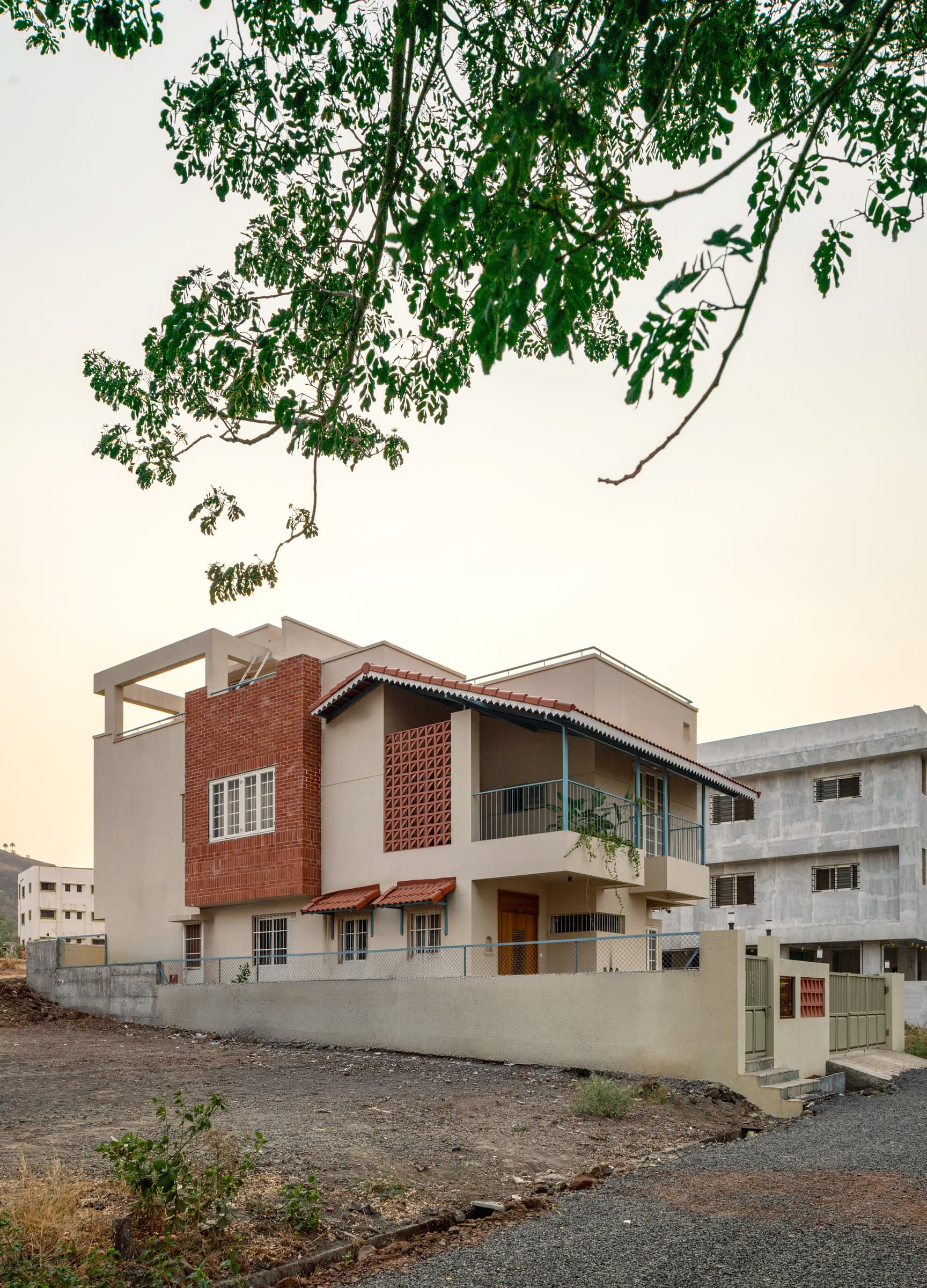 The street view at dusk: a cream stucco mass broken by a brick volume and a perforated terracotta jaali, with the Mangalore-tile awning establishing the regional register