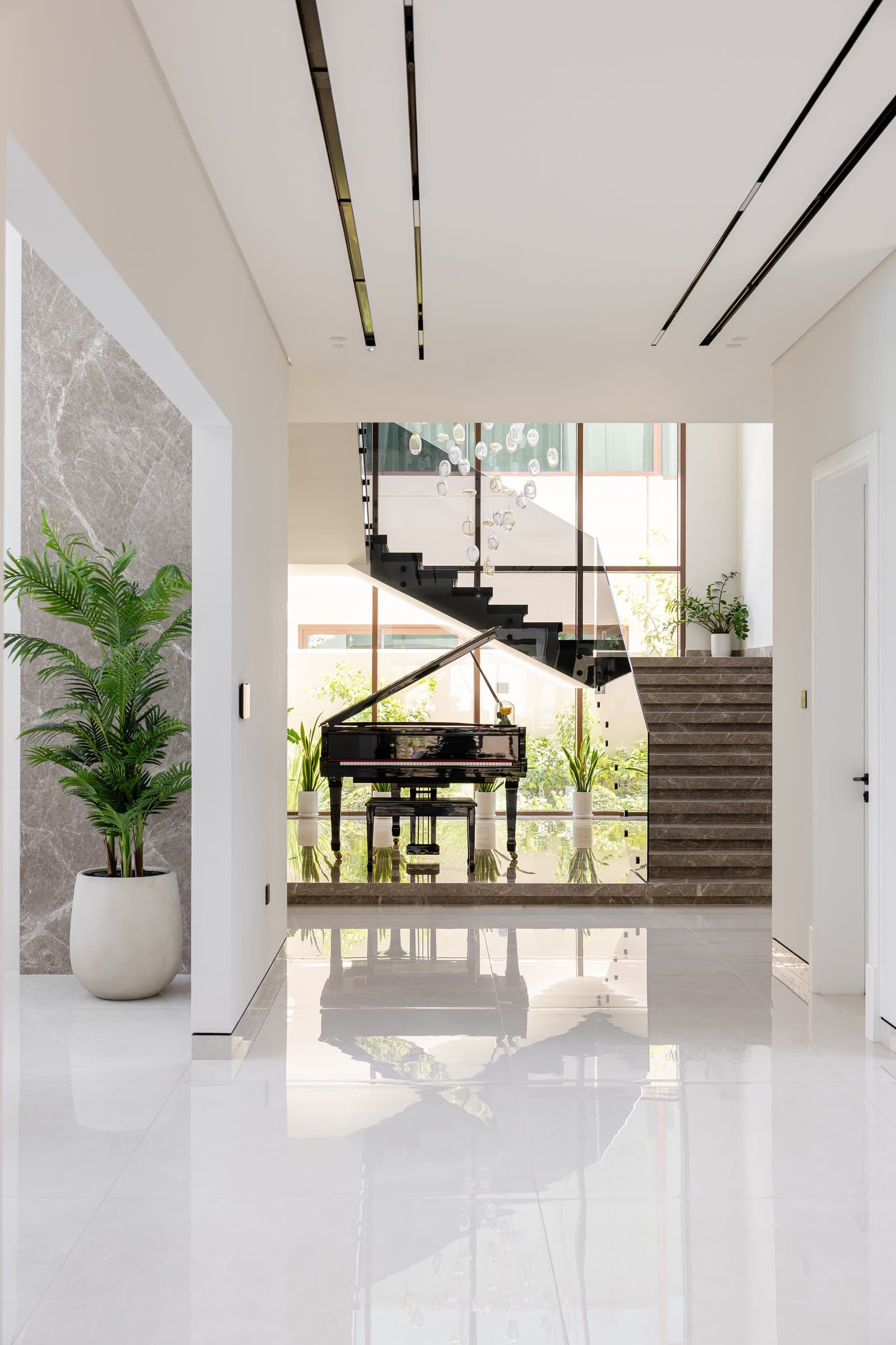 The arrival sequence, where a stone-clad portal frames the staircase, grand piano, and suspended glass installation rising through the double-volume void