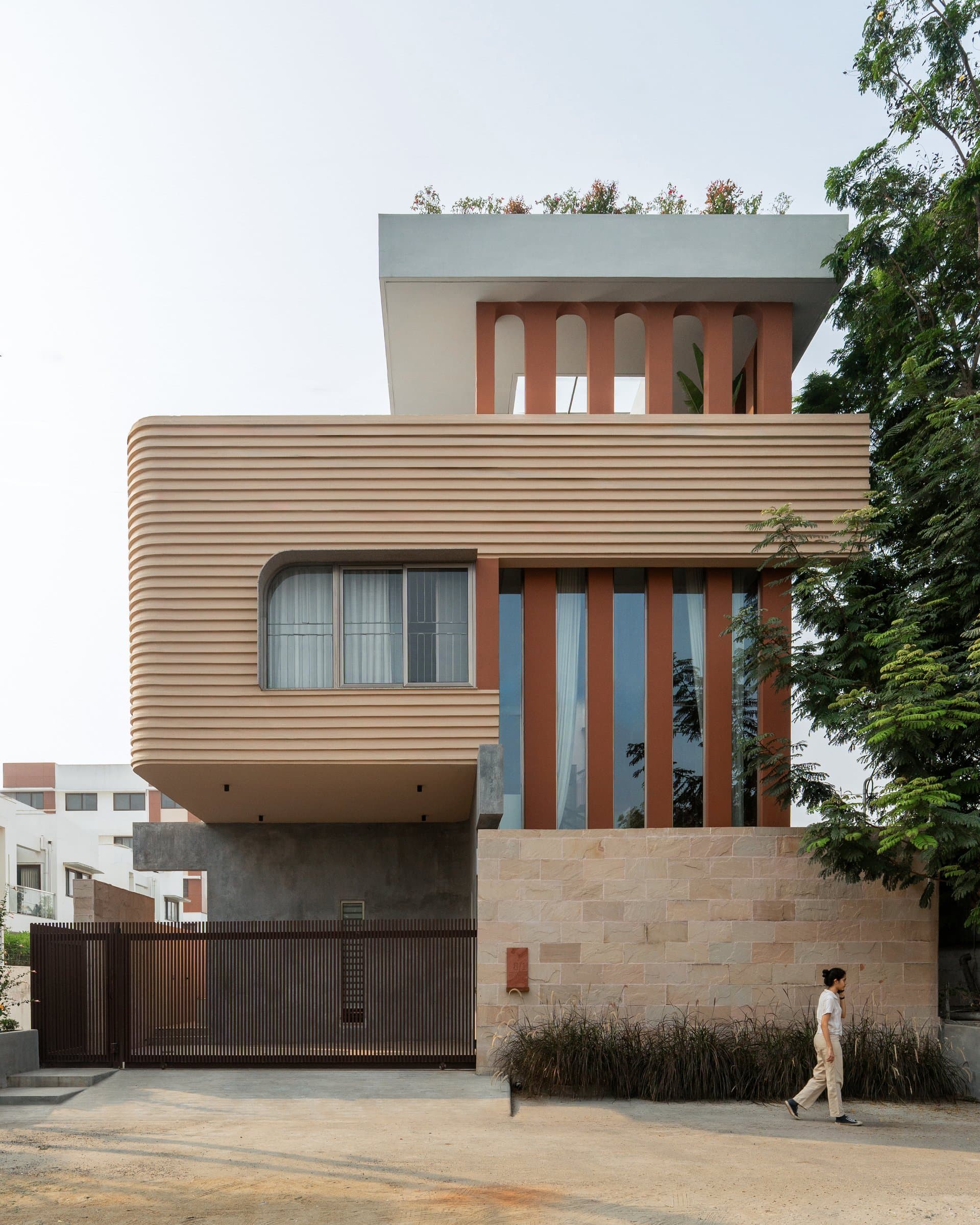 The street elevation, where a ribbed sandstone-toned volume cantilevers over a stone base beside a taller terracotta-finned volume
