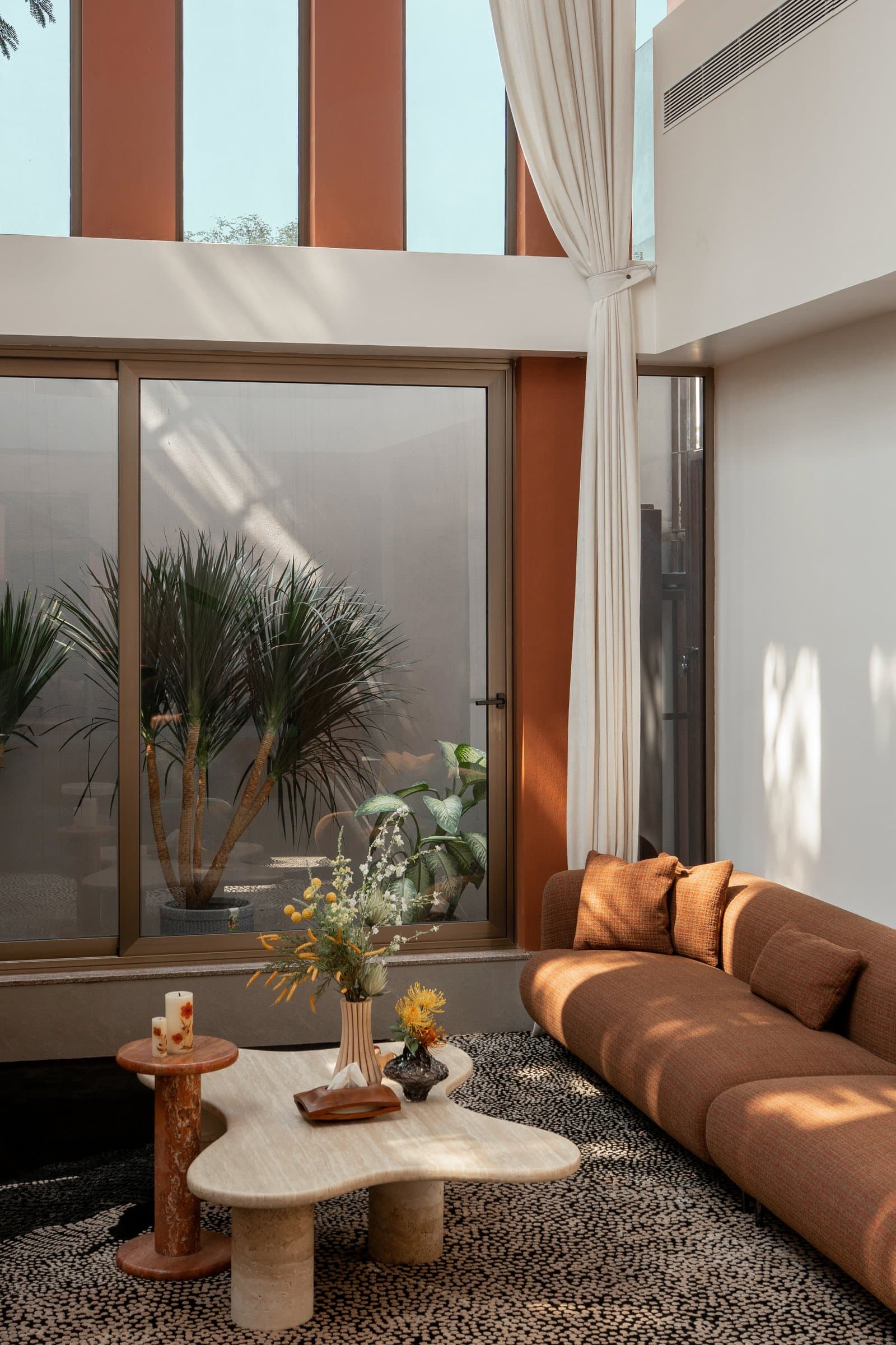 A quieter corner of the living space, where a rust-toned curved sofa opens onto a planted pocket courtyard through full-height glazing