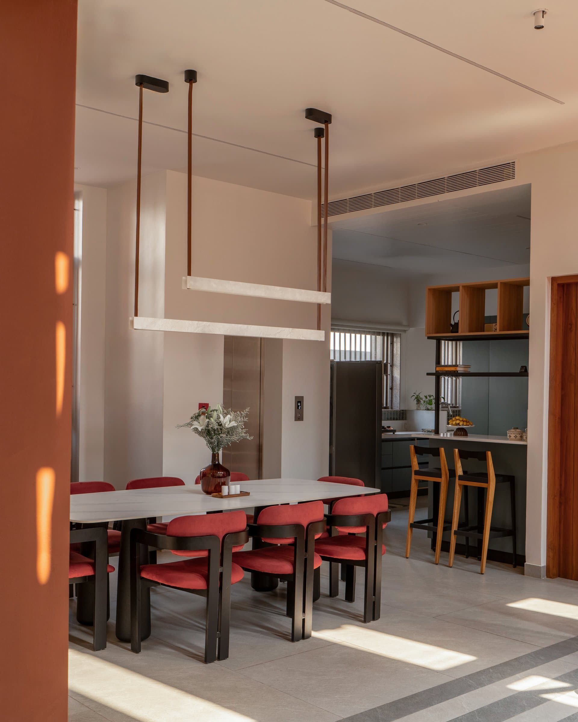 The dining area, where a stone-topped table and red-upholstered chairs sit under twin linear pendants beside a compact bar counter