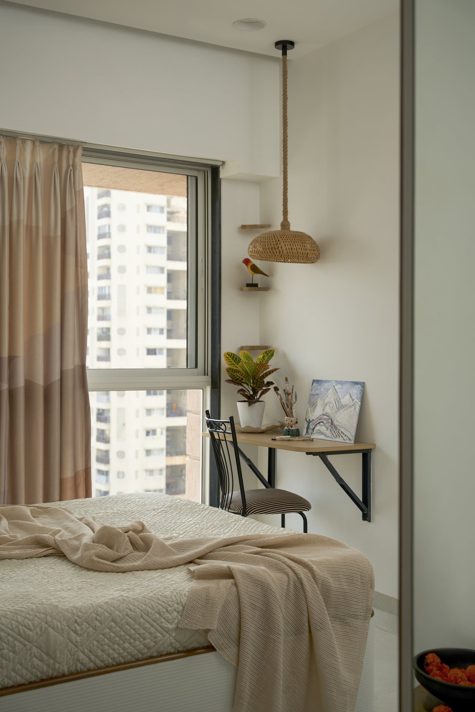 A small work corner tucked beside the window, with a wall-mounted timber shelf, a woven pendant on rope, and a slim corner shelf