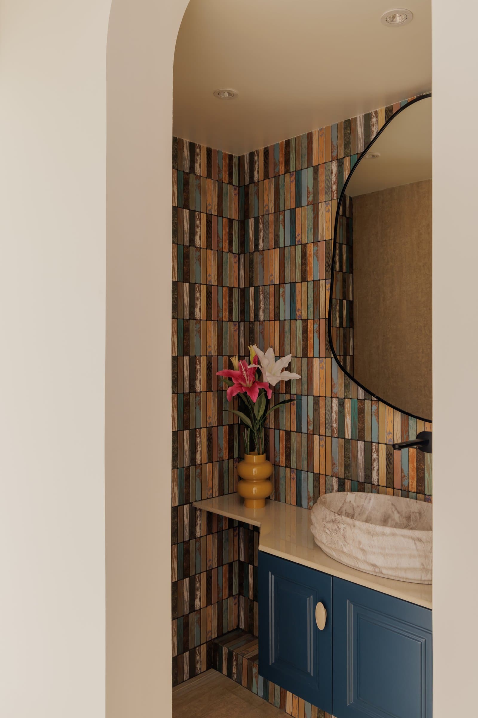 The powder room, where vertically stacked tiles in muted tones frame a sculpted stone basin and deep blue vanity