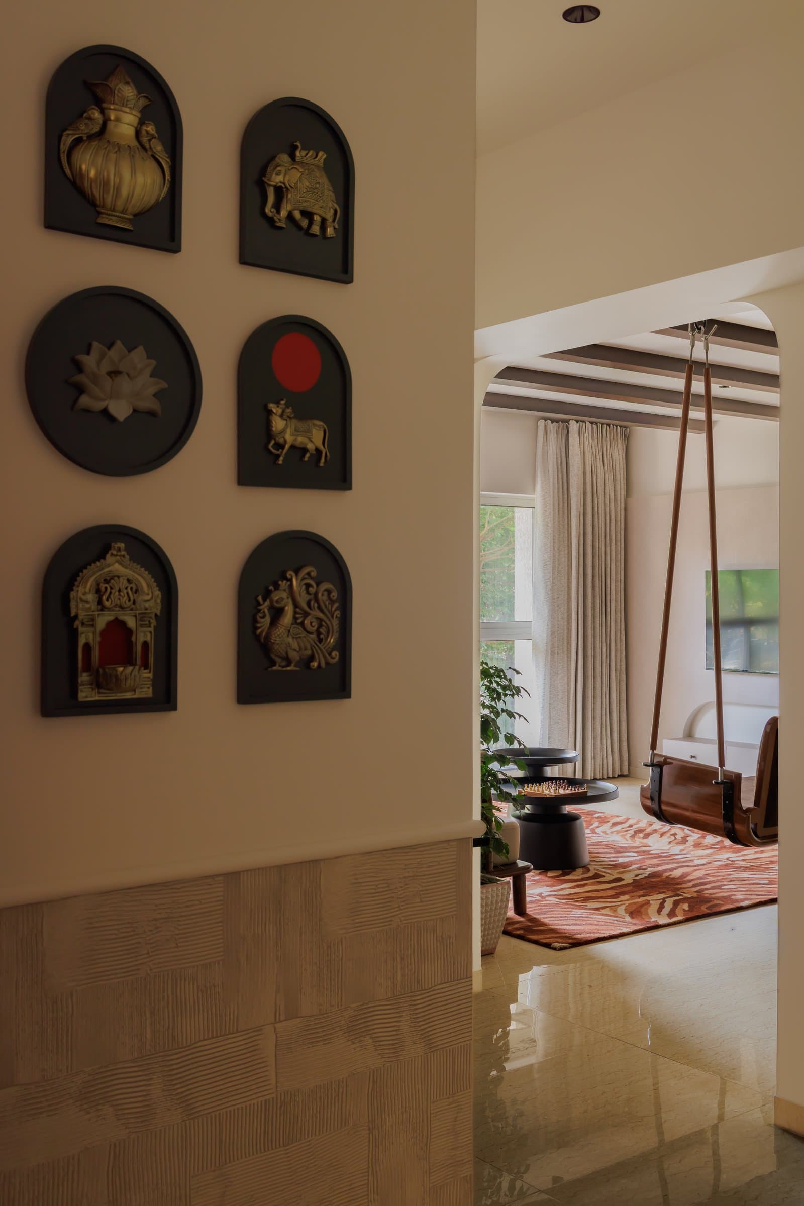 The passage wall, where arched brass reliefs of elephants, lotus, cow, and temple motifs frame a view into the living room