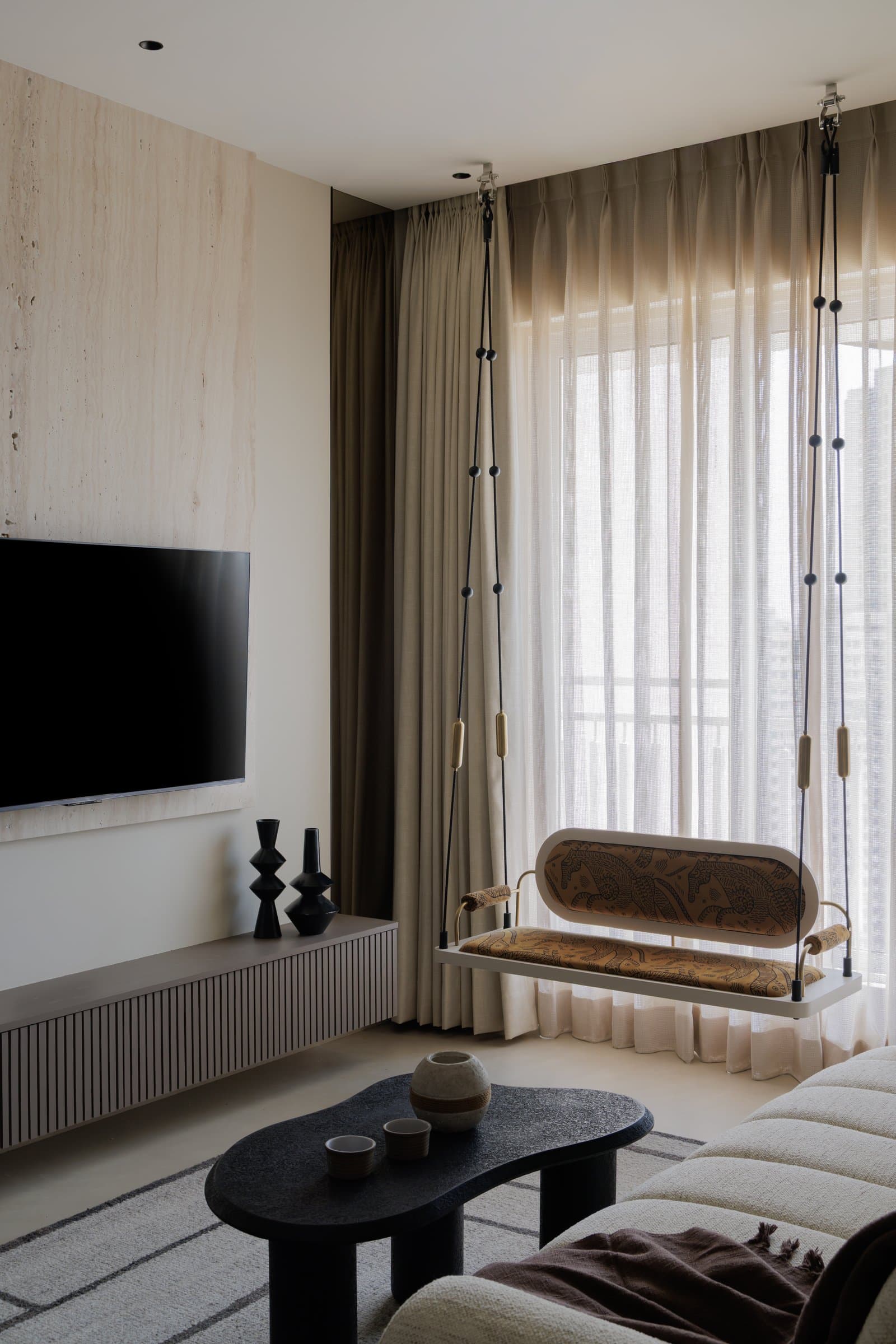 The media wall, where travertine and a fluted console anchor the television beside a beaded-rope swing at the window