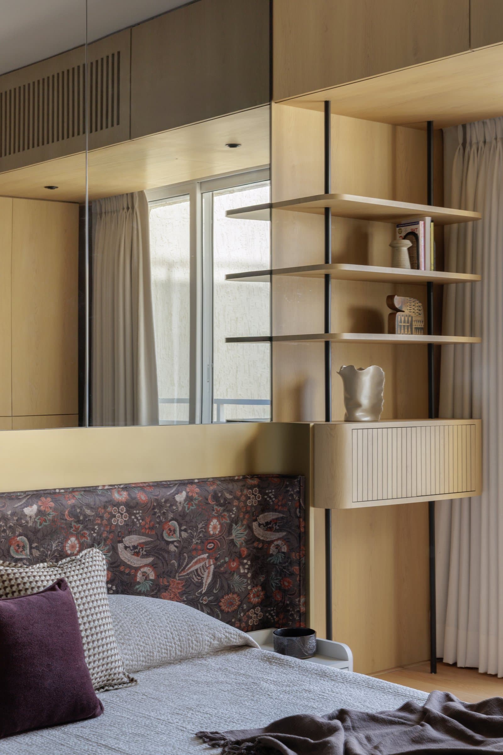 The master bedroom, where a densely printed floral headboard is framed by a brushed brass shelf and pale oak shelving