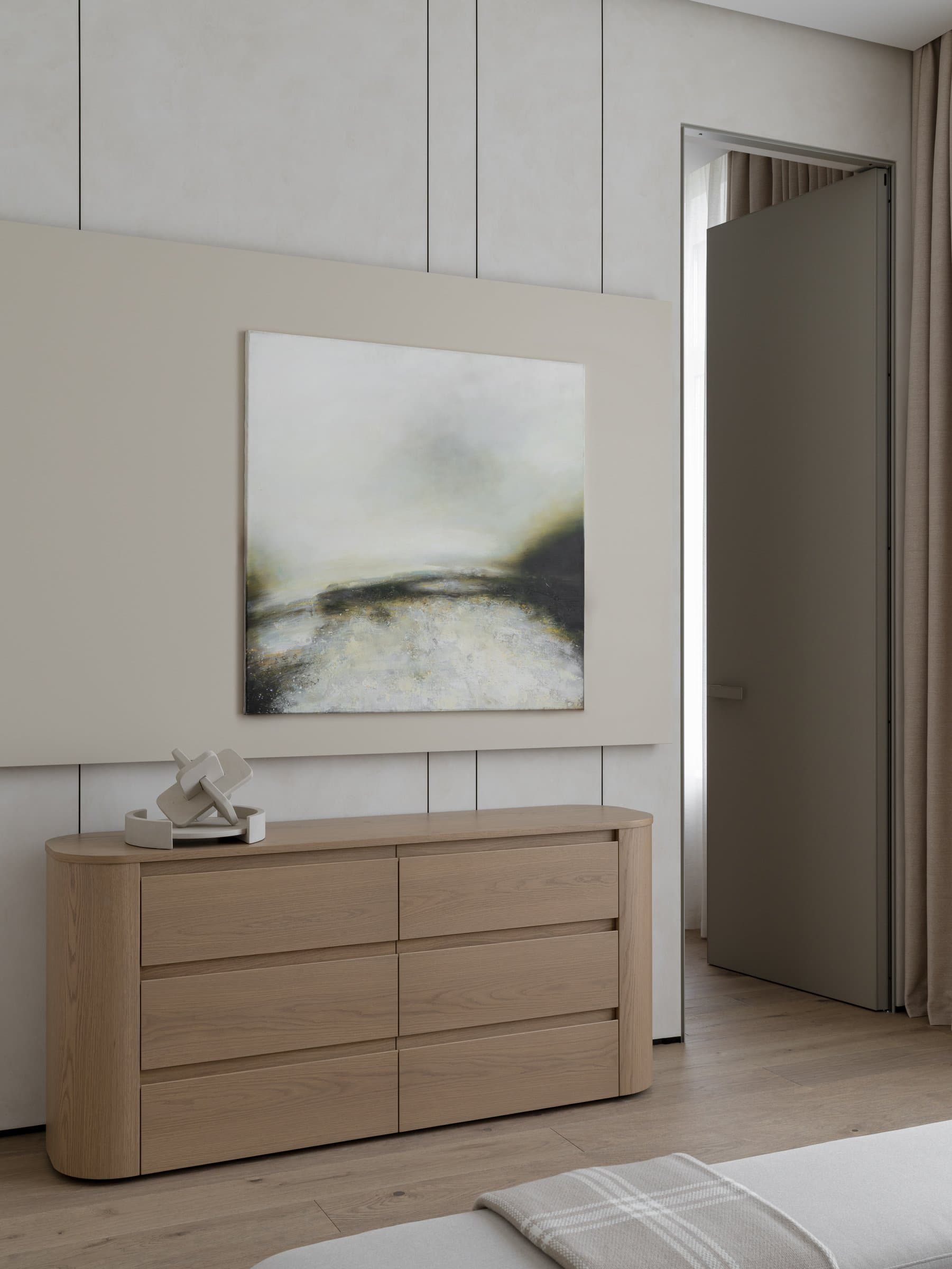 The chest of drawers with its curved edges, sitting below a horizon-line abstract painting in misty greens and greys