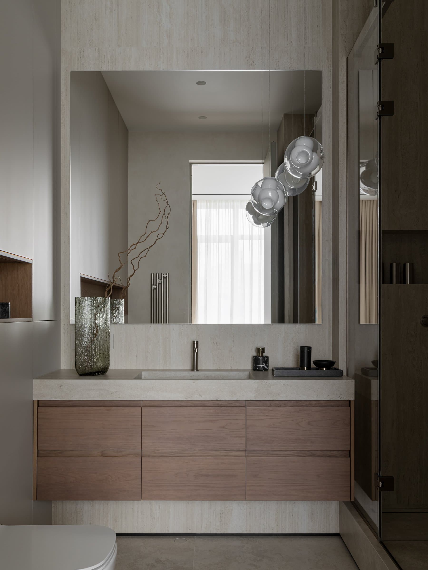 The primary bathroom, where a floating oak vanity meets travertine-toned stone and a cluster of glass sphere pendants