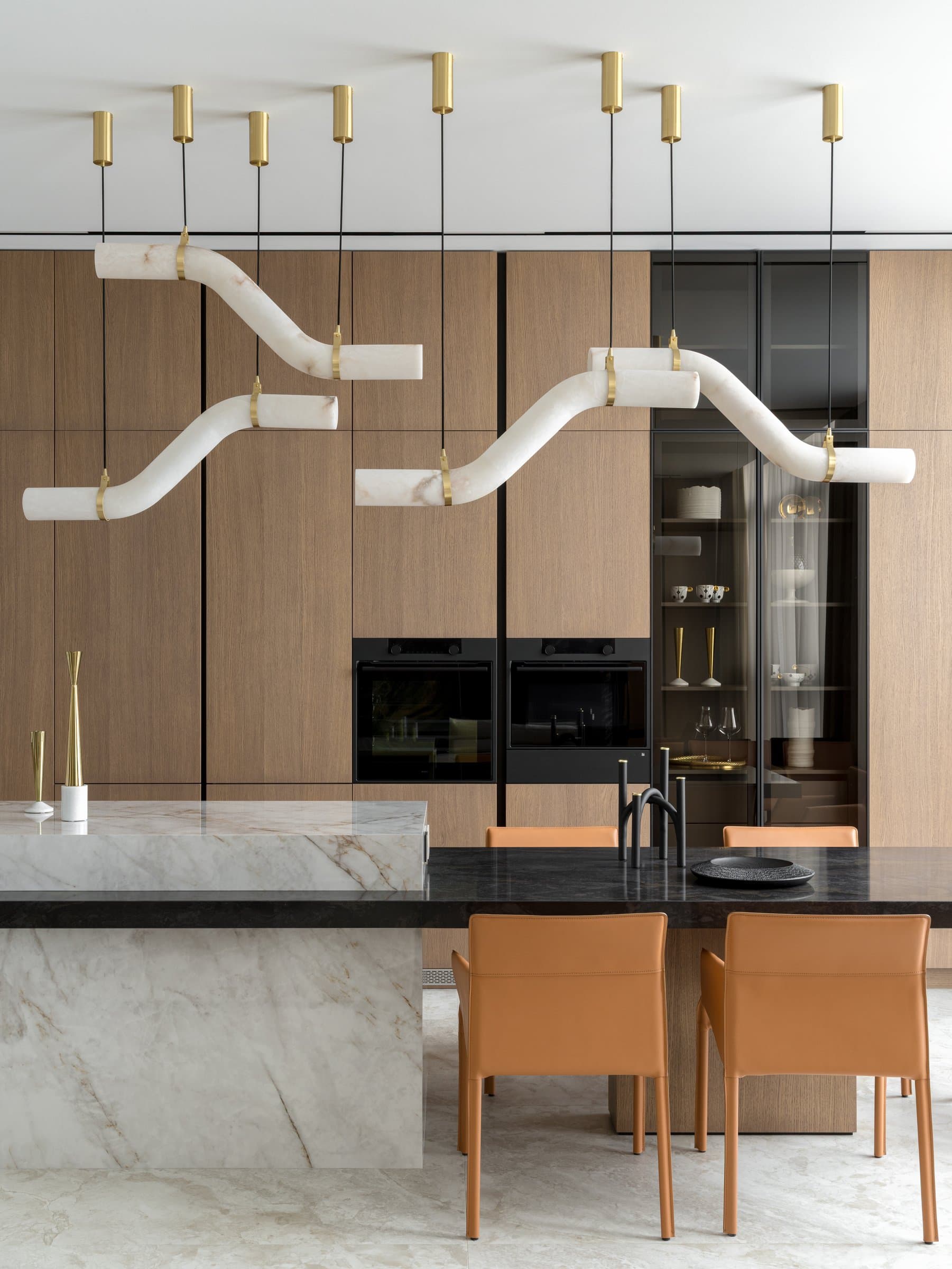 The pendant composition above the island: pale stone tubes suspended from slim brass ceiling mounts, bending in shallow S-curves