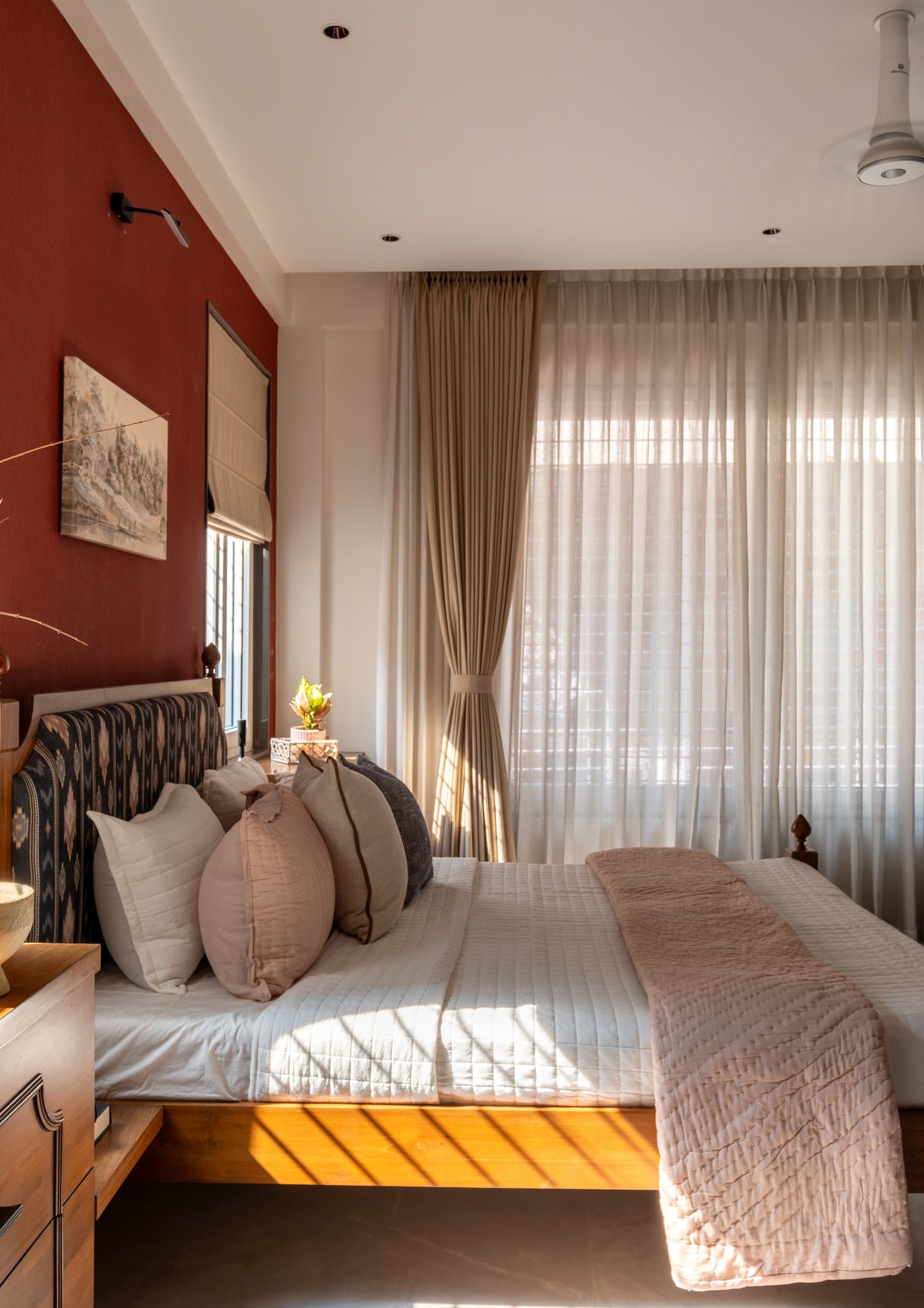 The bed, dressed in muted neutrals and ikat cushions, with a red accent wall carrying a small artwork
