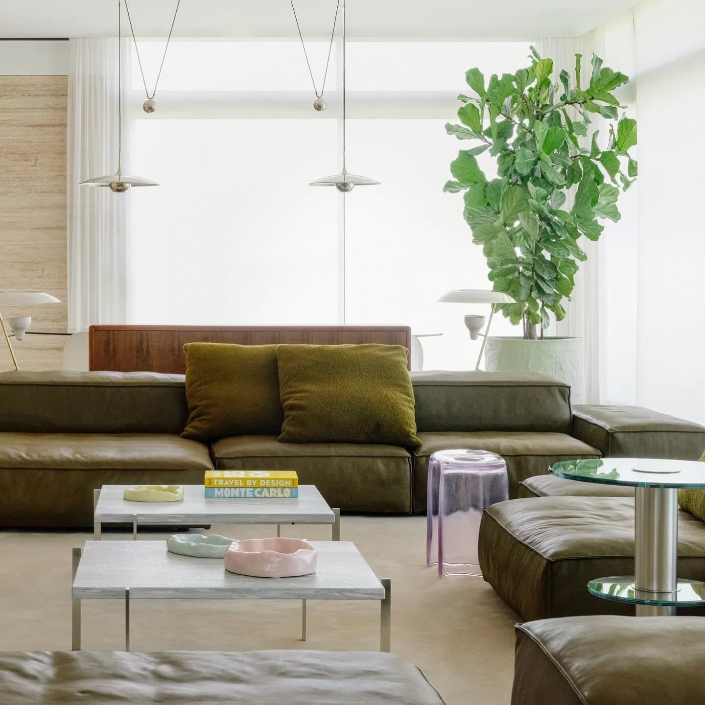 The sunken living area gathers around low stone-topped tables, its olive leather sofas grounding a pale, airy composition.