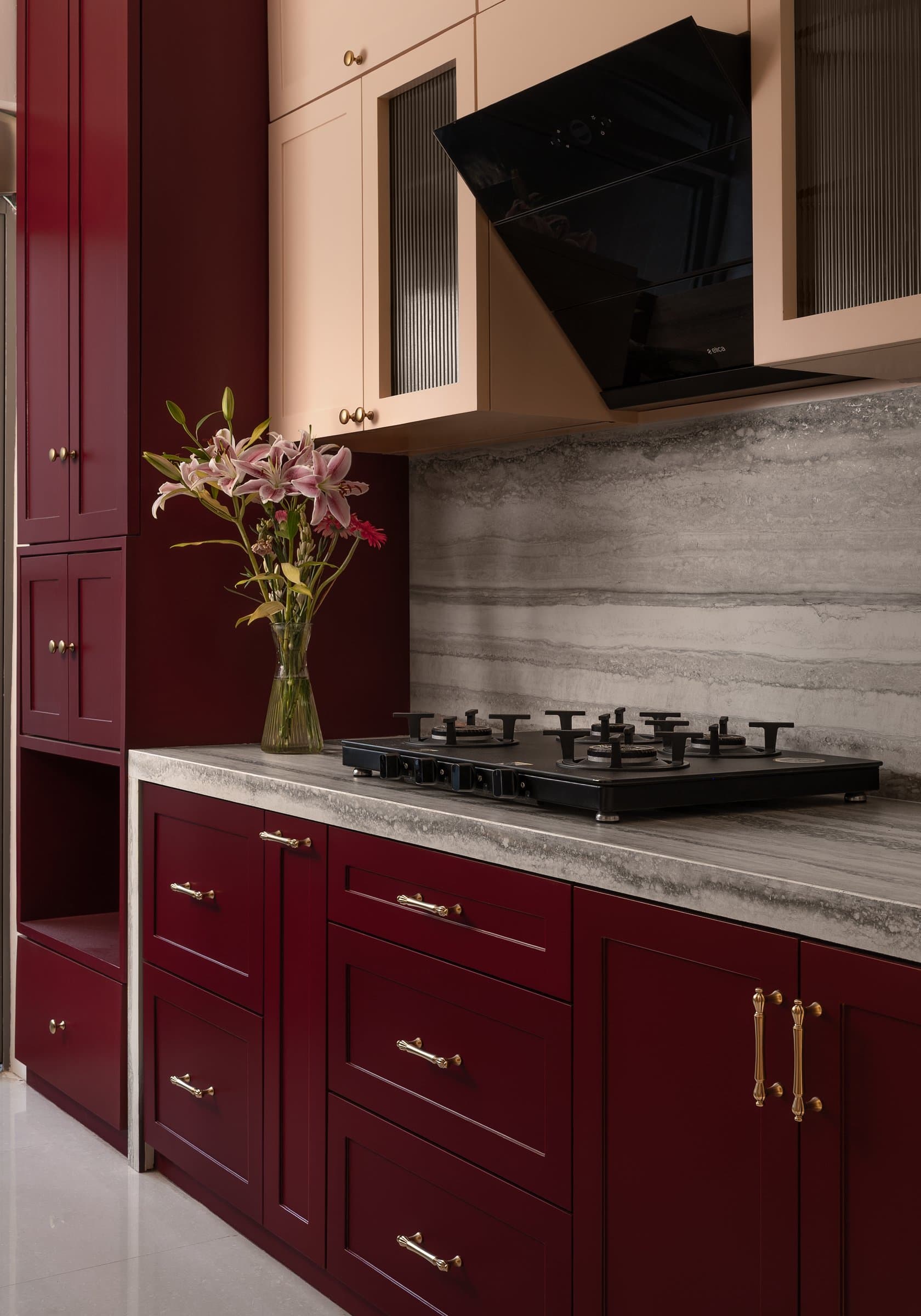 The kitchen, where deep oxblood cabinetry with brass pulls meets soft blush uppers across a veined grey stone counter