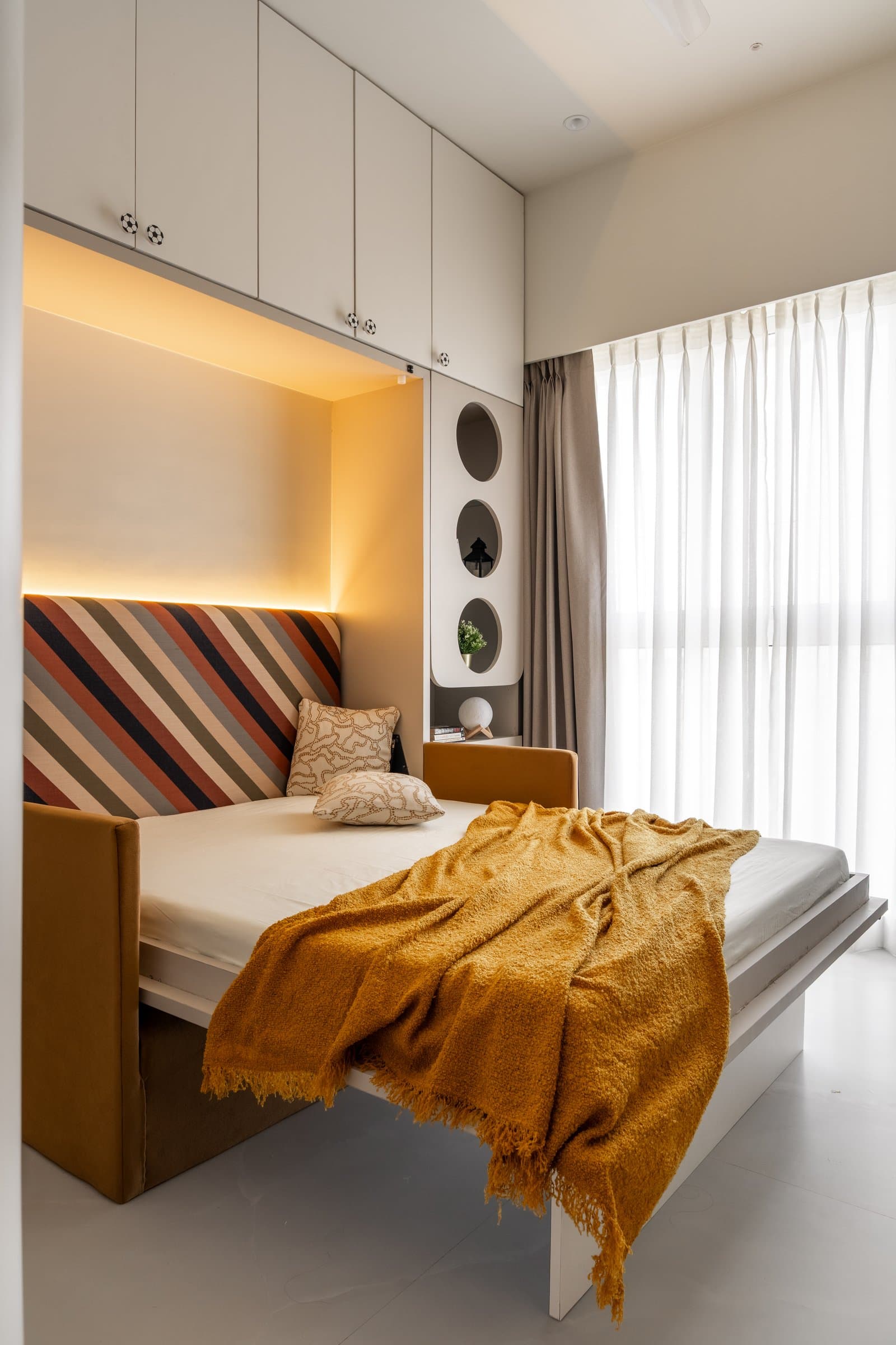 The son's bedroom, where a foldaway bed with a striped headboard tucks into a cove-lit niche, freeing the floor for daytime use